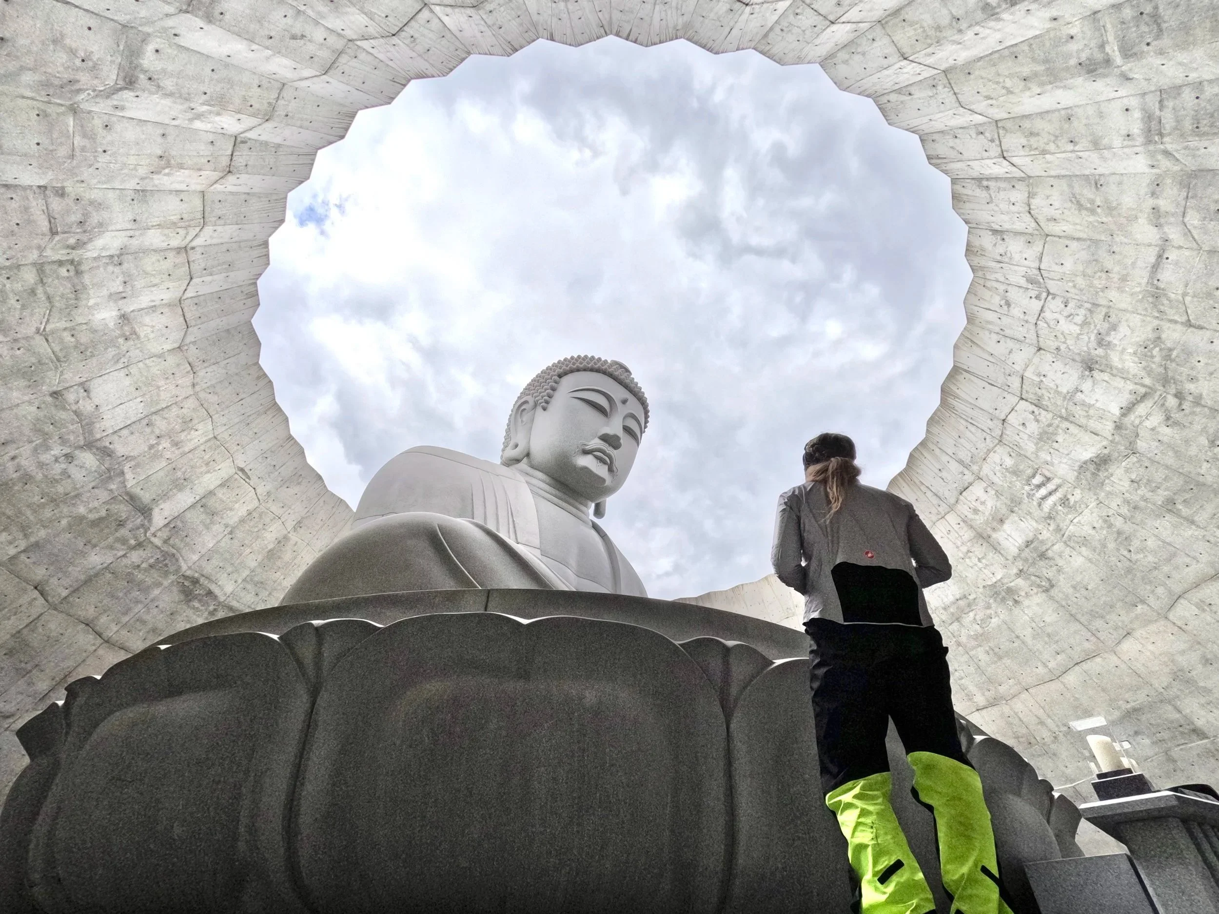 A person standing inside a circular opening in the ceiling, looking up at a large outdoor Buddha statue. The Buddha is carved from stone and is positioned against a cloudy sky.