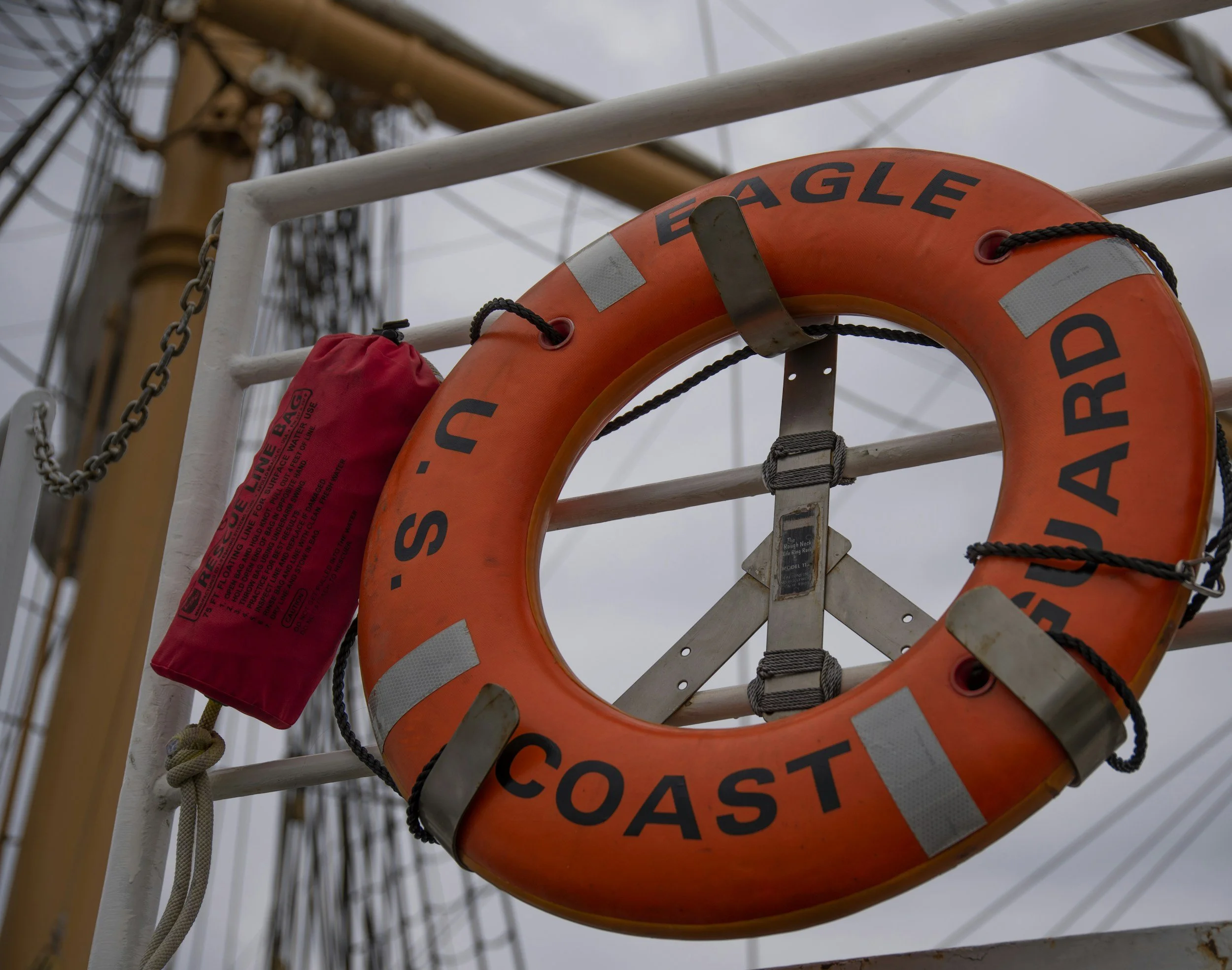 An orange lifebuoy with the words "U.S. EAGLE COAST GUARD" written on it, attached to a white railing on a ship, with a red rescue bag hanging nearby and a ship's mast with rigging and wires in the background.