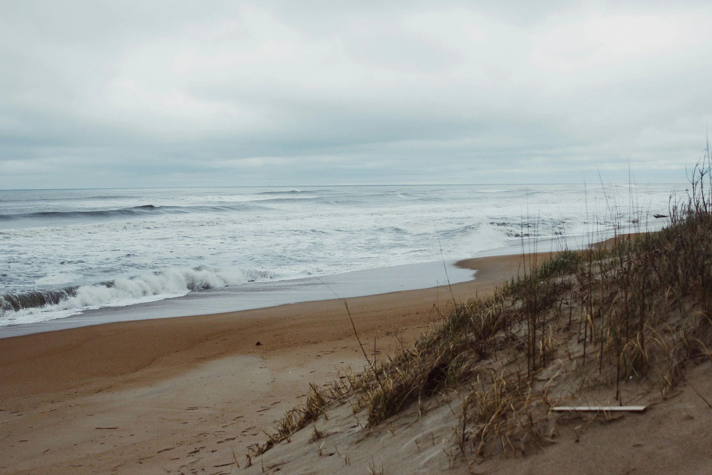Overcast beach scene with sandy dunes, sparse grass, and ocean waves