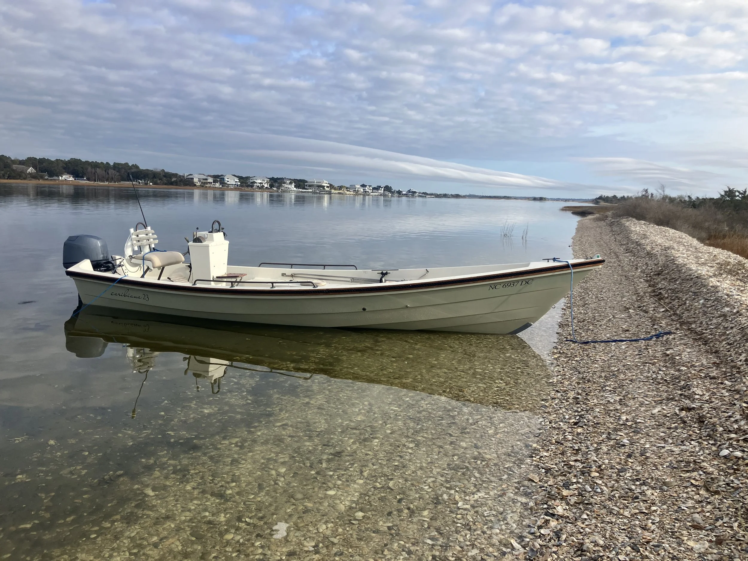 A small white motorboat with beige seats is docked on a pebbly shore in Swansboro, with its reflection visible in the calm water. In the distance, there are houses along the shoreline under a cloudy sky.