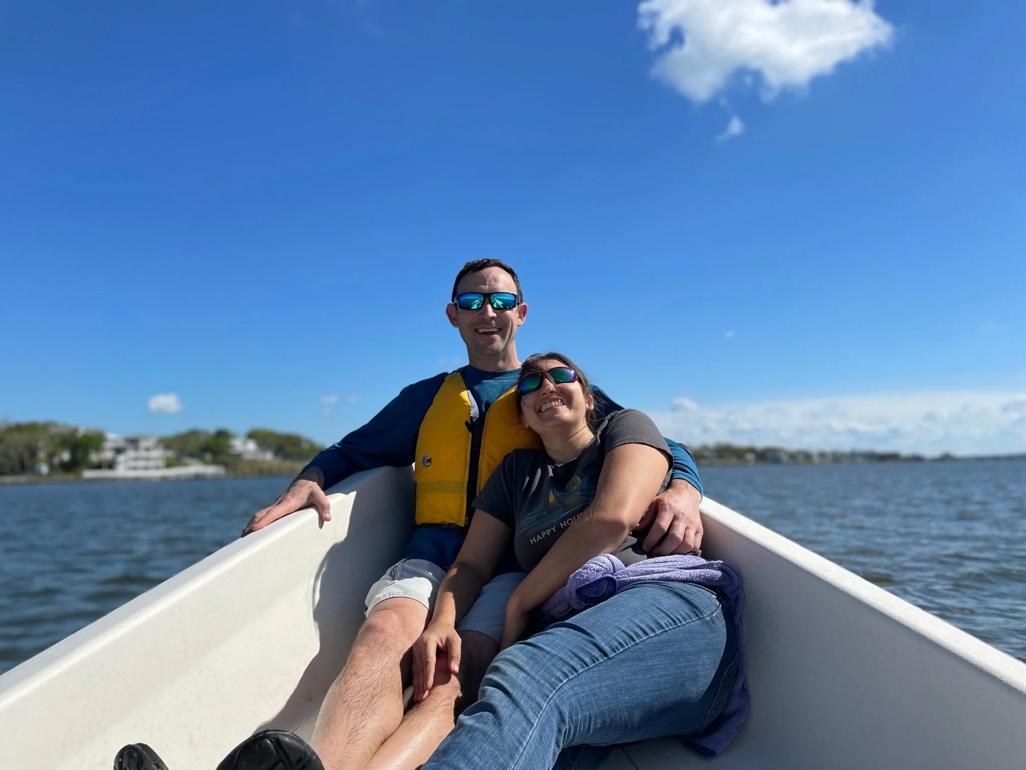 A smiling man and woman sitting close together in a boat, enjoying a sunny day on the water with a shoreline and houses in the background.