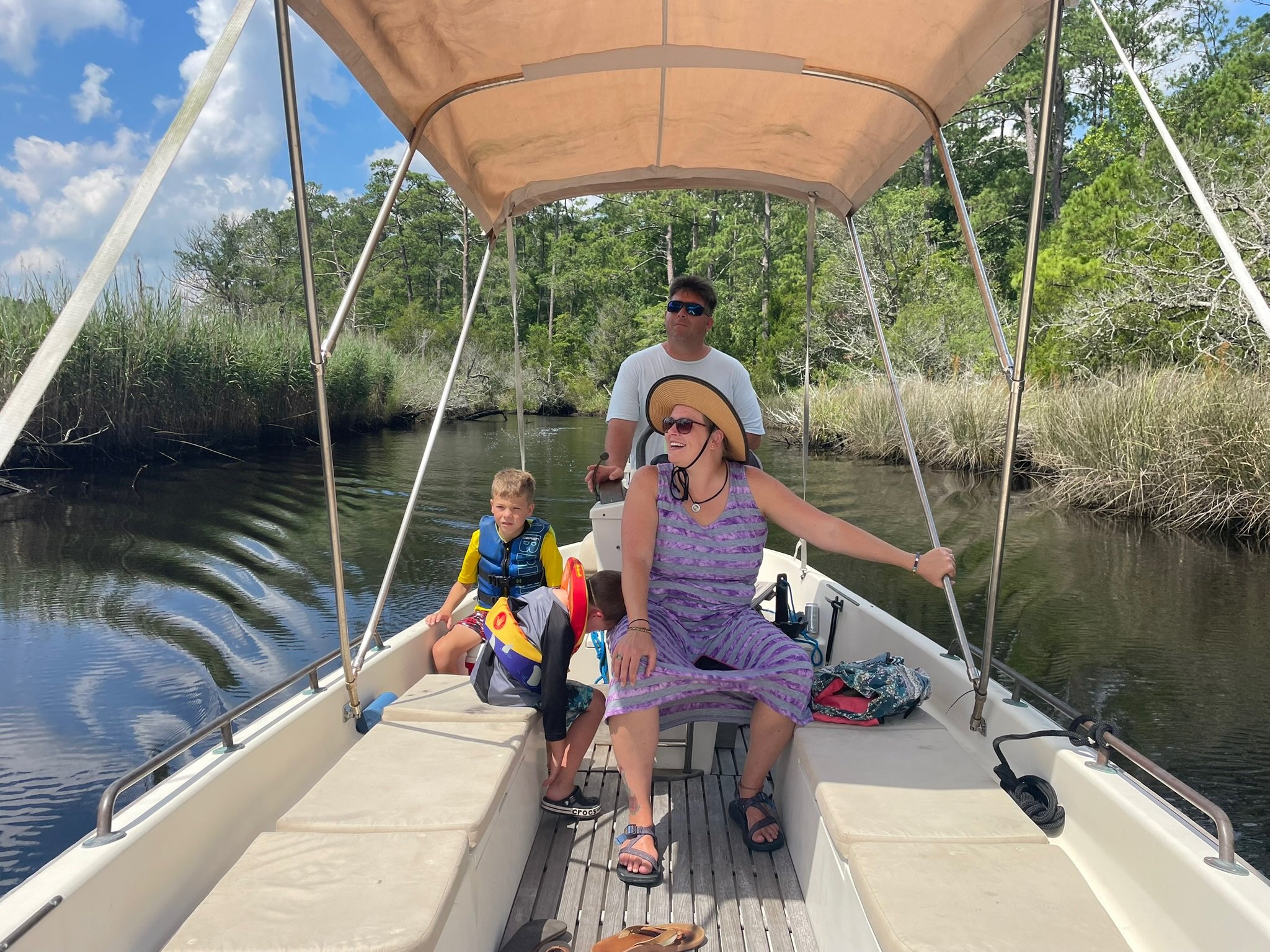 A family sailing on a boat through Swansboro's White Oak River surrounded by lush green trees and grass, with three people on the boat enjoying the day.