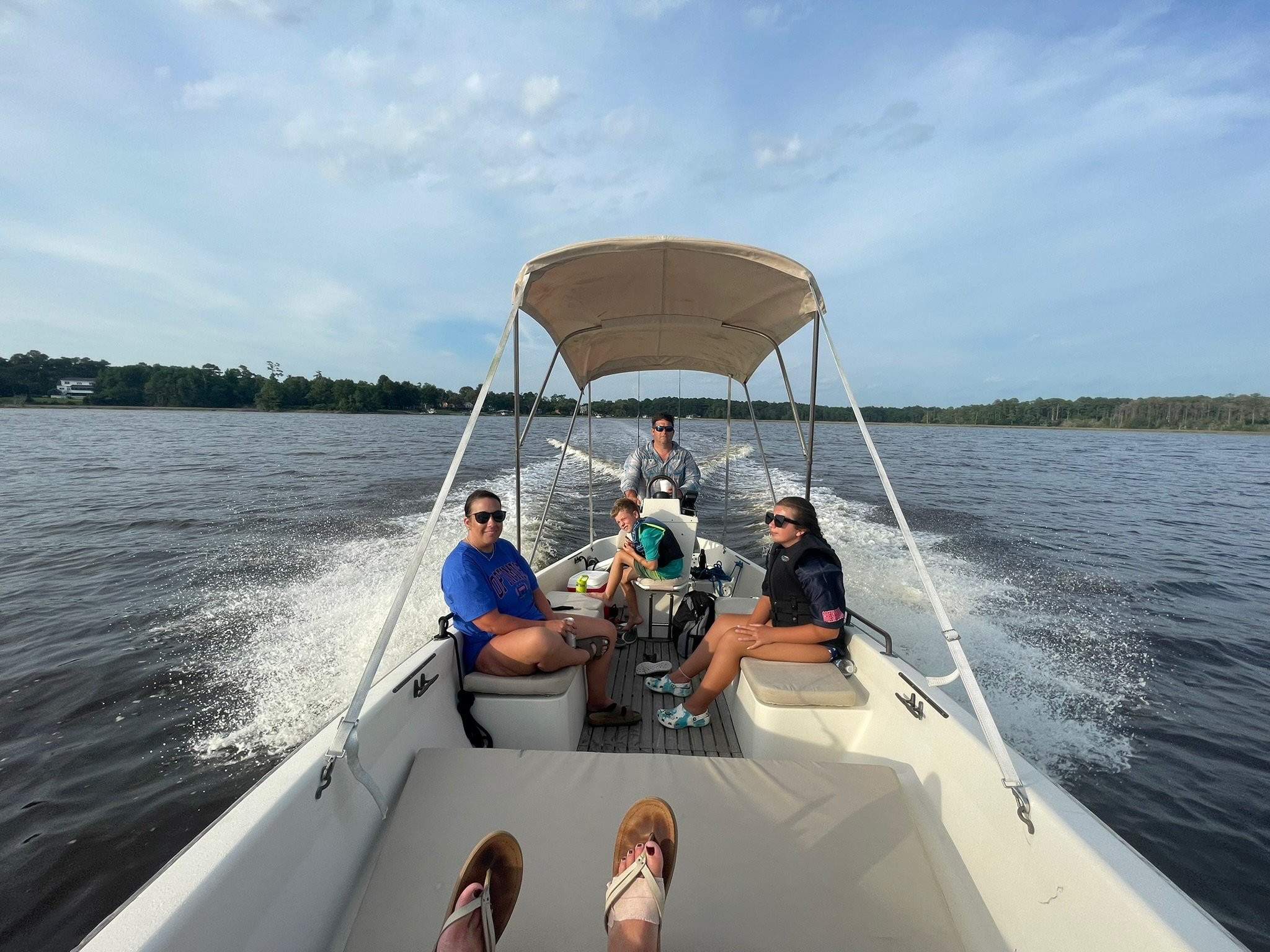 Four people, including a young girl, are on a boat riding on a large lake with trees in the background and a partly cloudy sky.