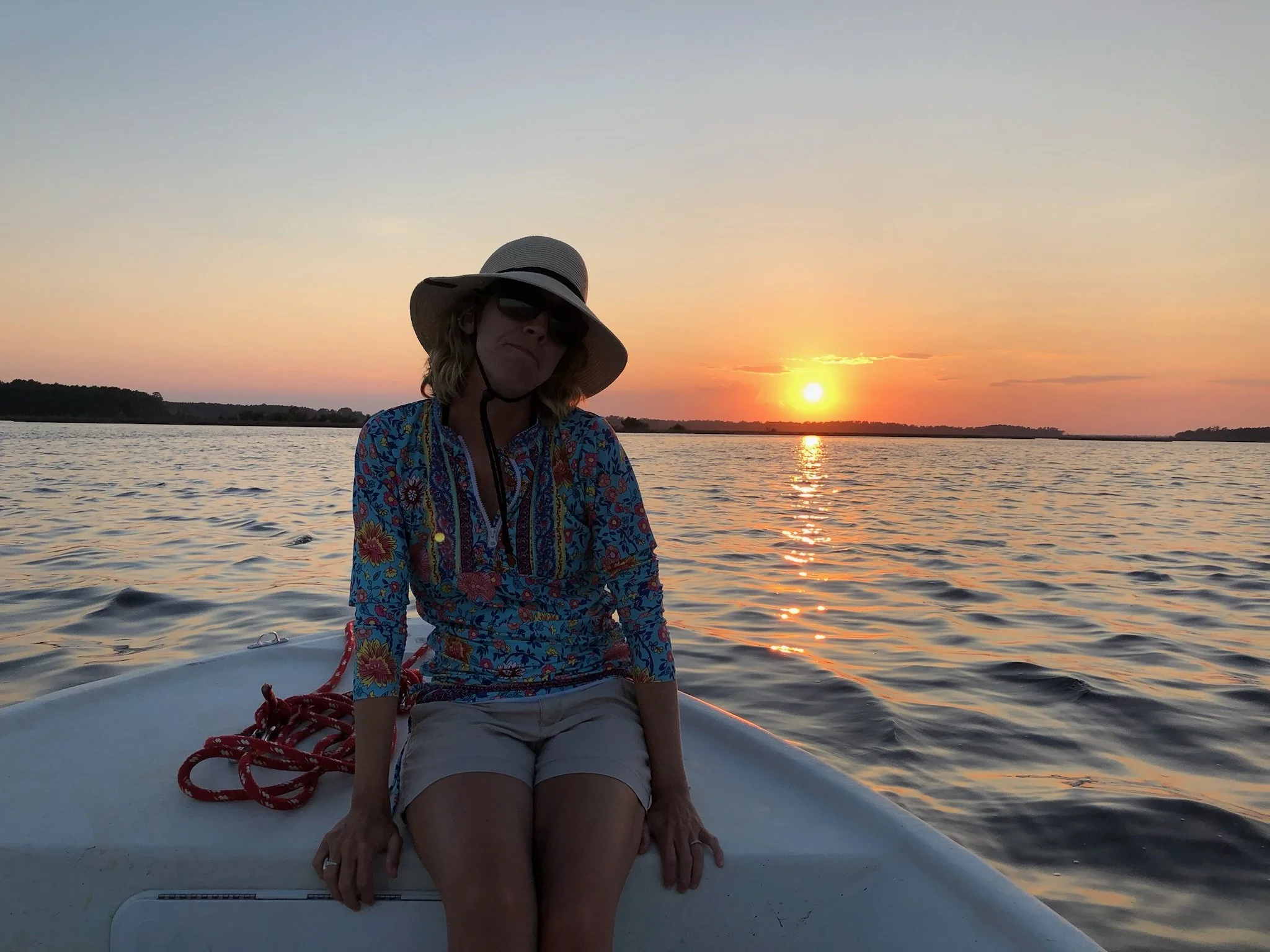 Woman sitting on the edge of a boat during sunset, wearing a wide-brimmed hat, sunglasses, a colorful floral top, and beige shorts, with water and a sunset in the background.