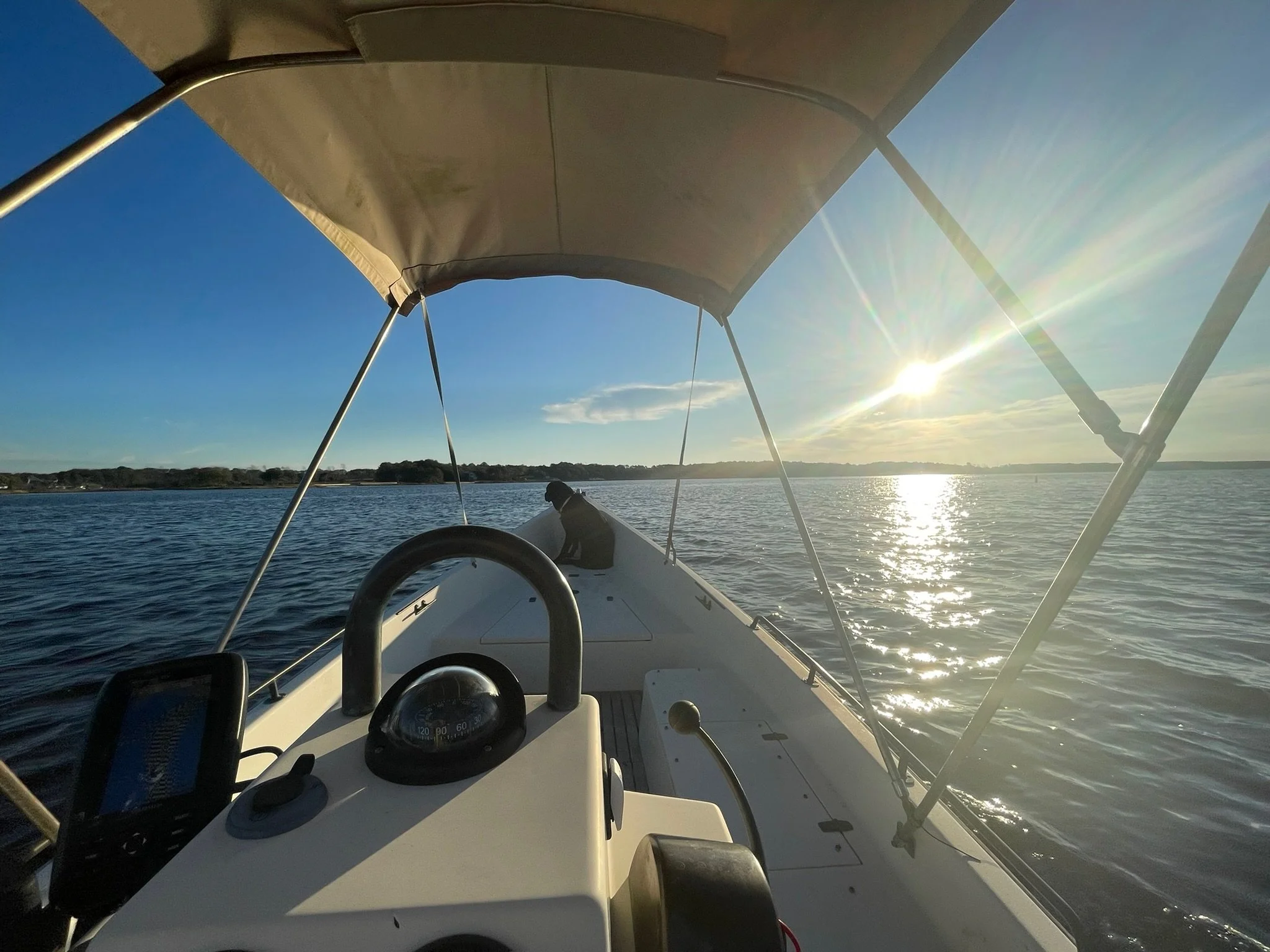 View from a boat showing the steering console and a person sitting at the bow, with the sun shining over a body of water during sunset or sunrise.