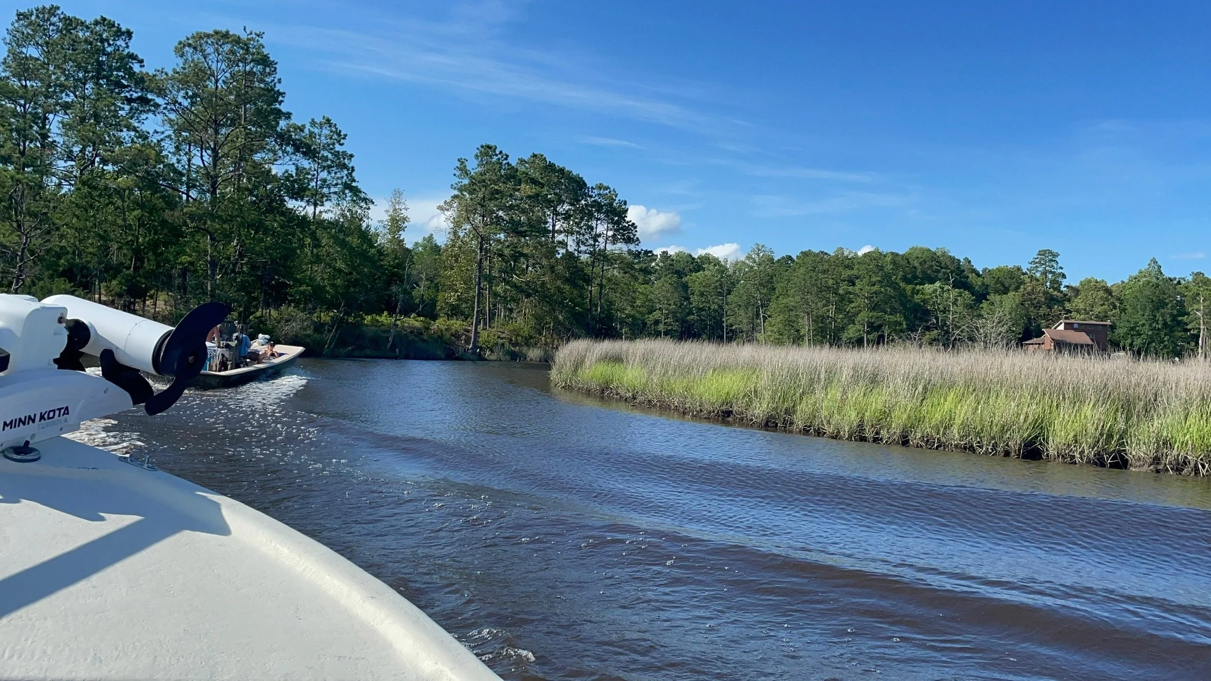 A boat moving through a river, with a motor visible in the foreground and a person on a second boat in the distance, surrounded by trees and reeds, under a blue sky.