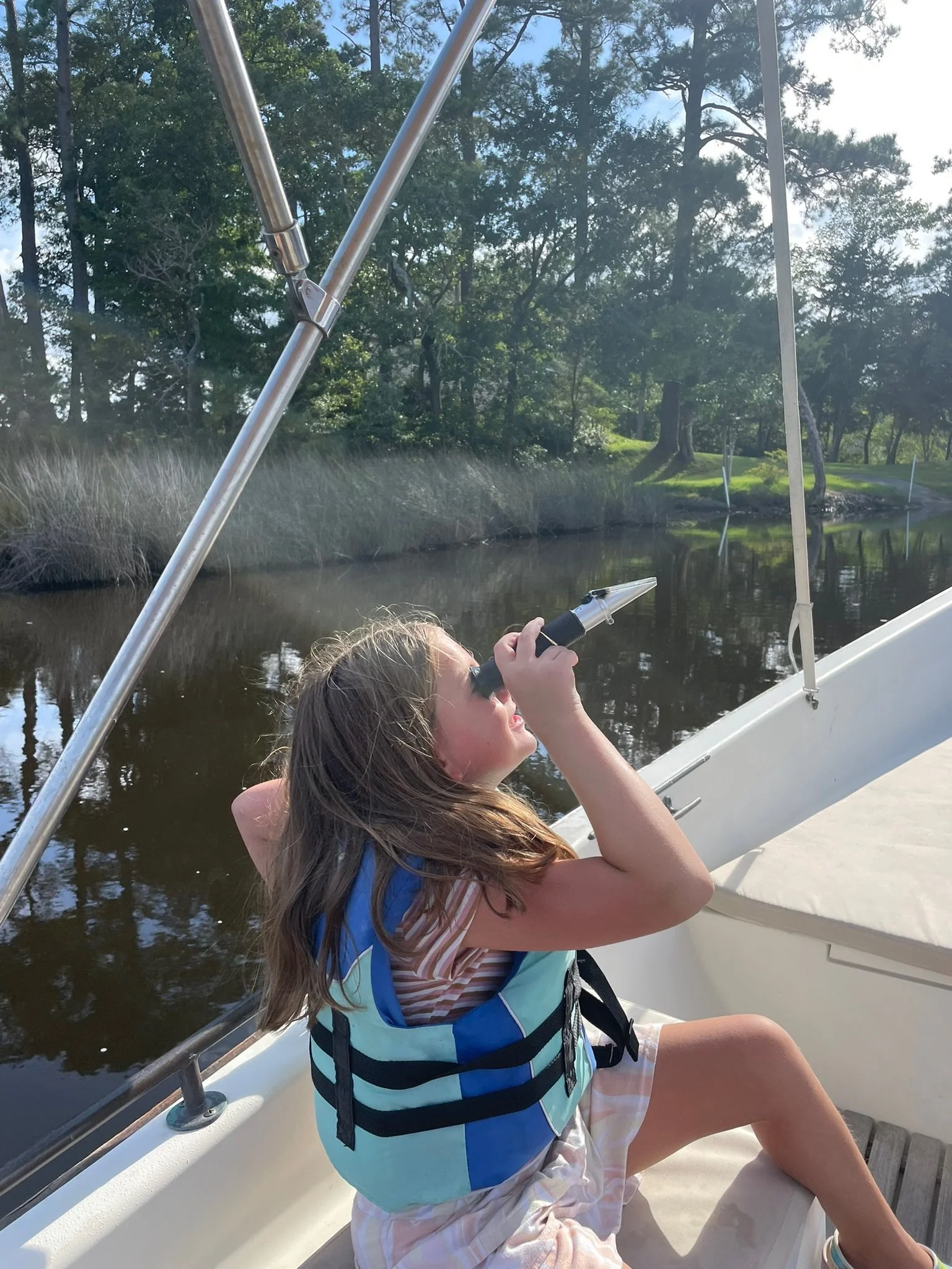 A young girl on a boat, sitting near the edge, looking through a telescope, with a river and green trees in the background.