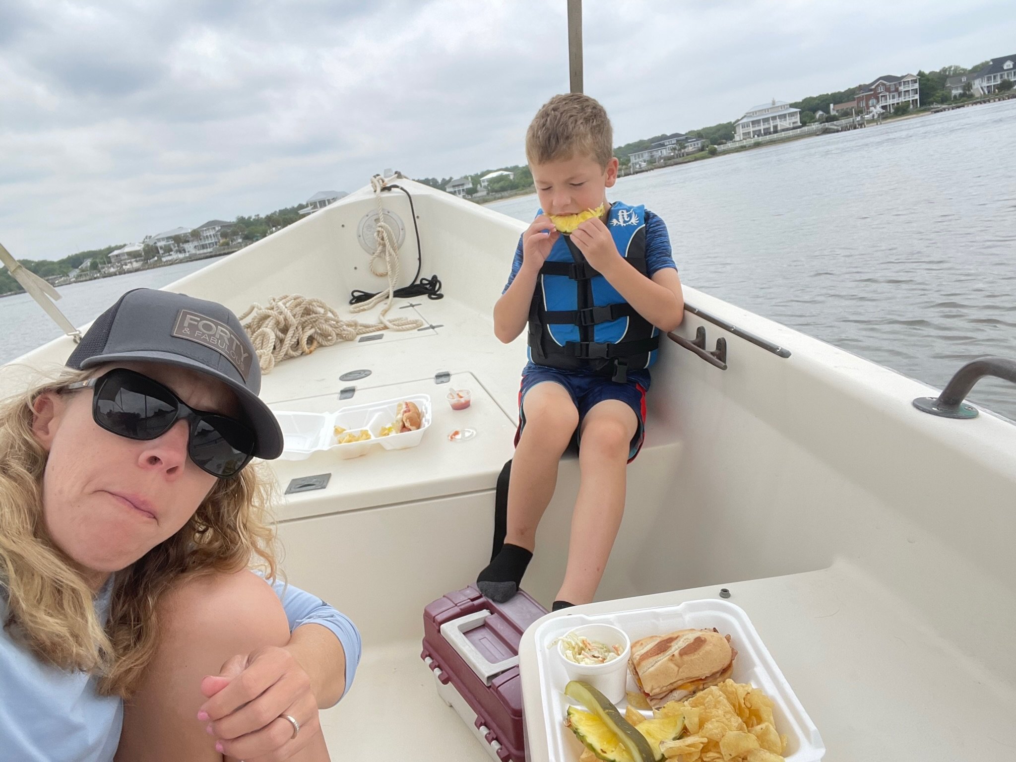 A woman and a boy on a boat, with the woman taking a selfie. The boy is eating a hot dog, and there is a tray of food with chips, coleslaw, and a pickle in front of him. The background shows water and houses along the shoreline under a cloudy sky.