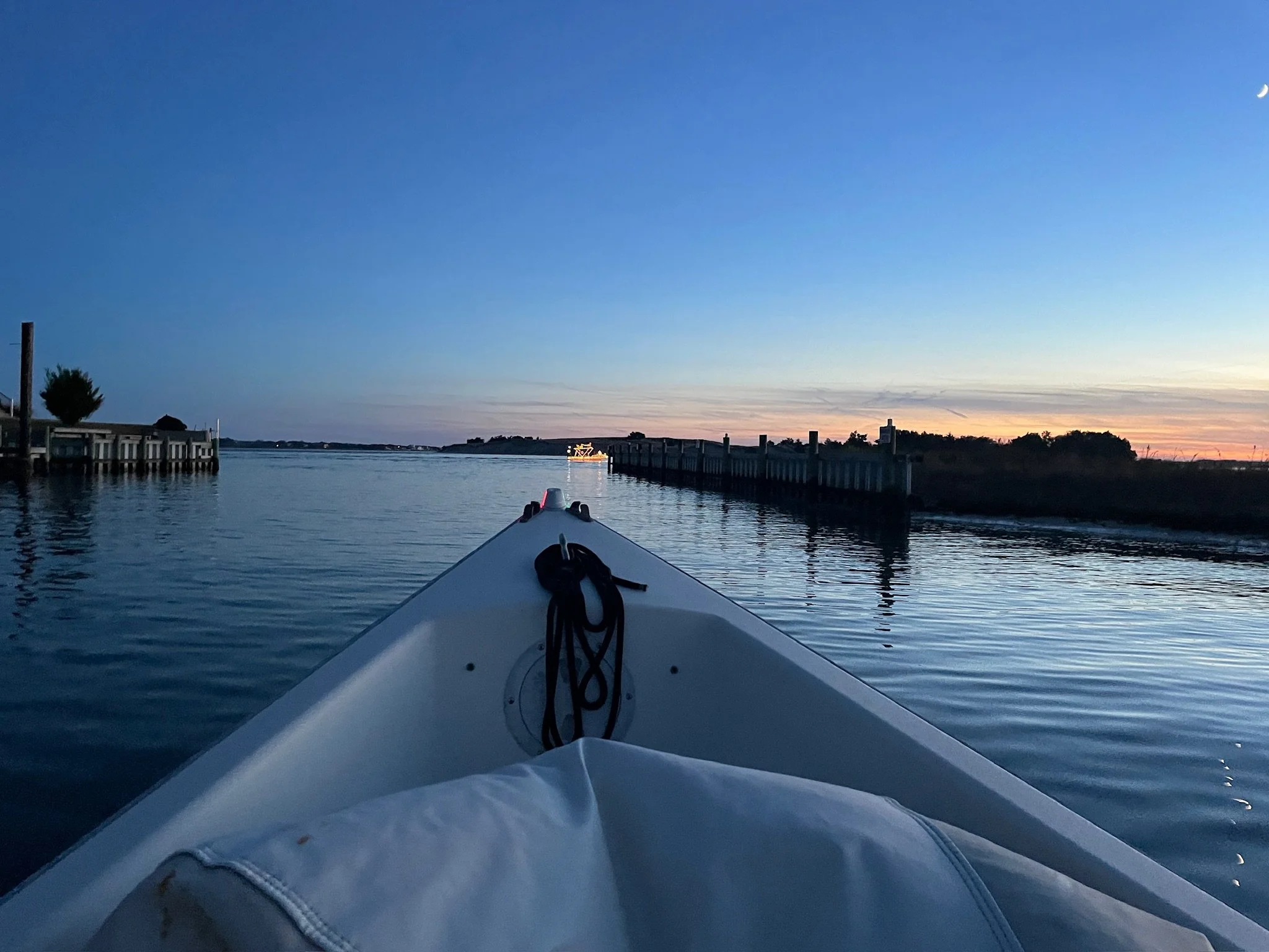 A boat on White Oak River,  during dusk, with docks on either side and a distant boat illuminated against the twilight sky.
