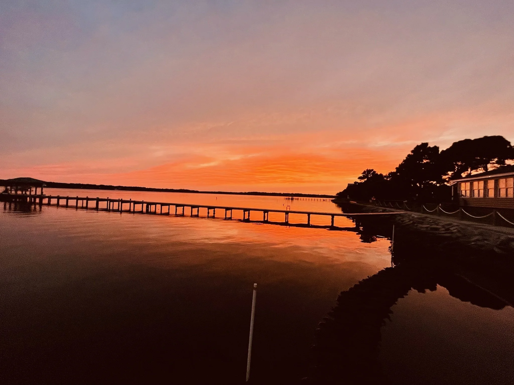 Sunset over a calm river with a pier extending into the water, silhouetted trees on the right, and buildings along the shore.