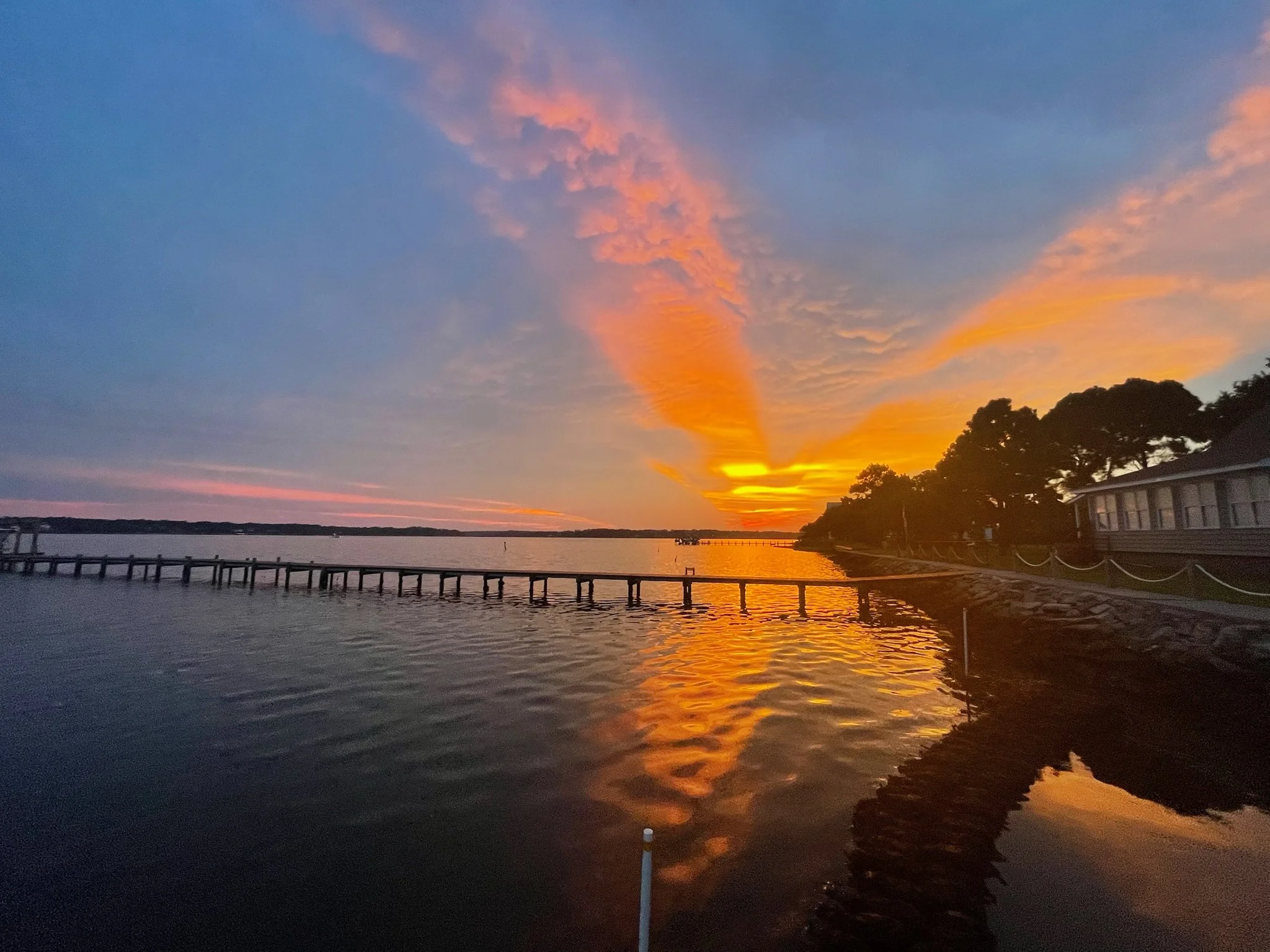 Sunset over a White Oak River with a dock extending into the water, silhouetted trees on the right, and a building on the far right, with vibrant orange, pink, and purple clouds in the sky.