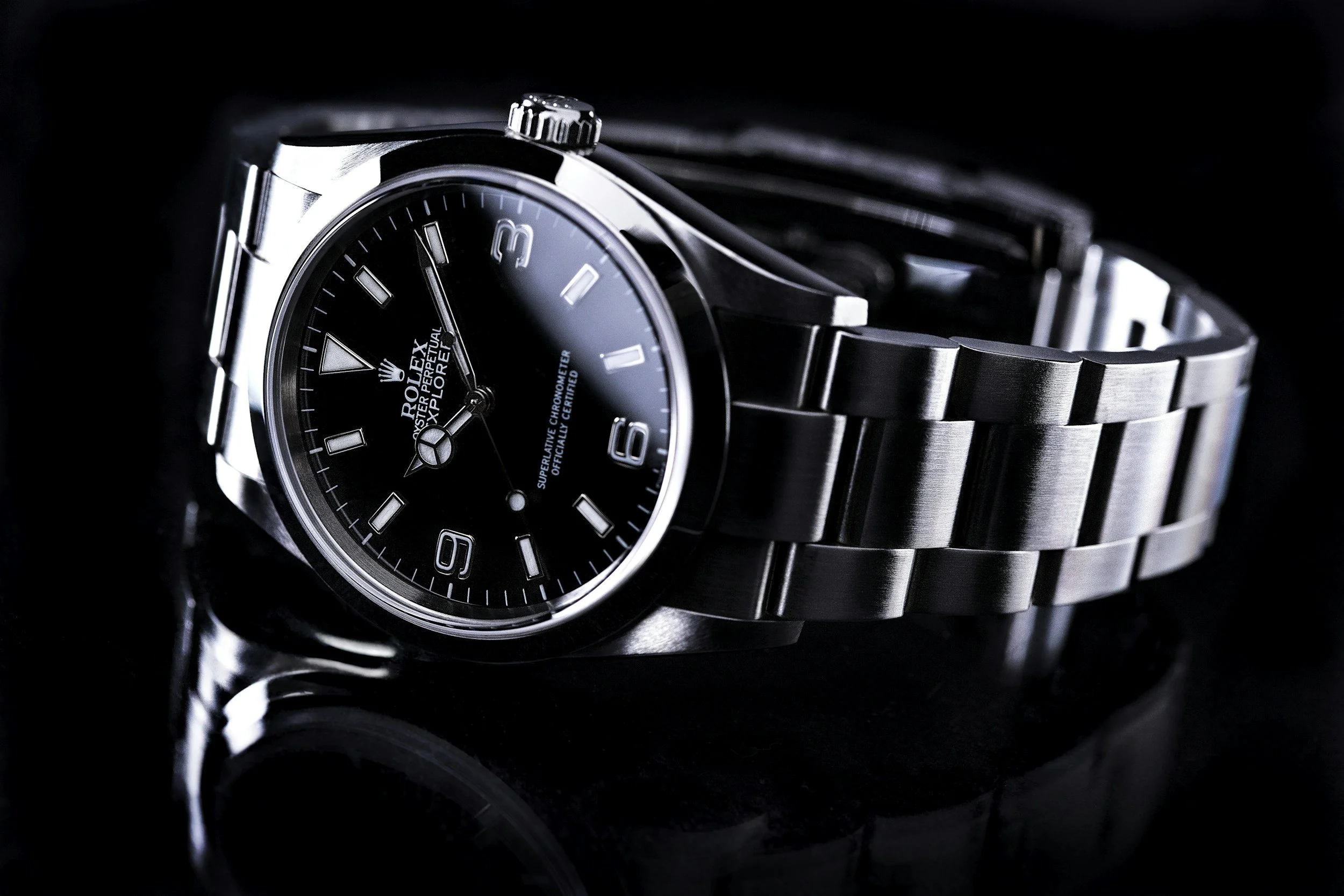 Close-up of a stainless steel Rolex Explorer watch with a black dial, silver markers, and bracelet on a dark reflective surface.