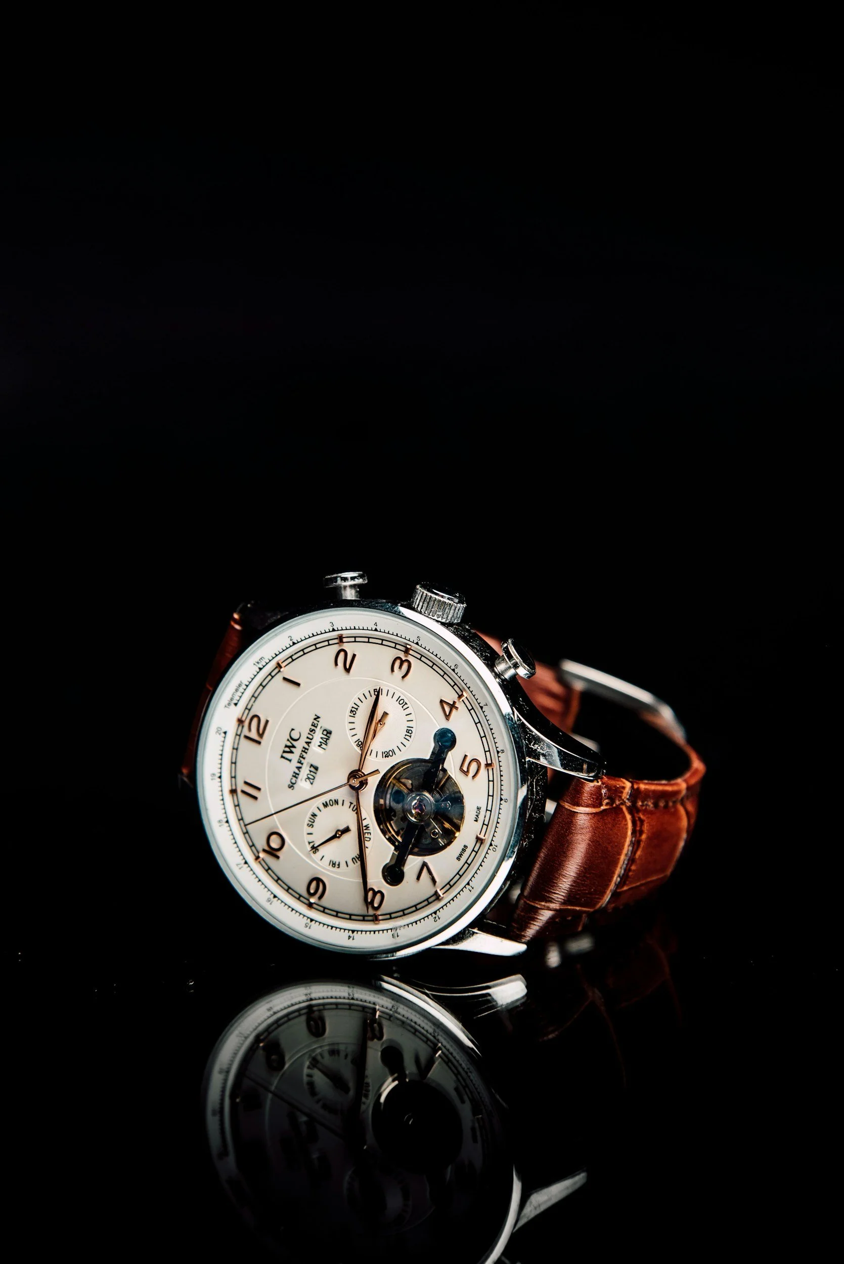 Luxury wristwatch with a white dial, multiple sub-dials, and a brown leather strap, reflected on a glossy black surface.