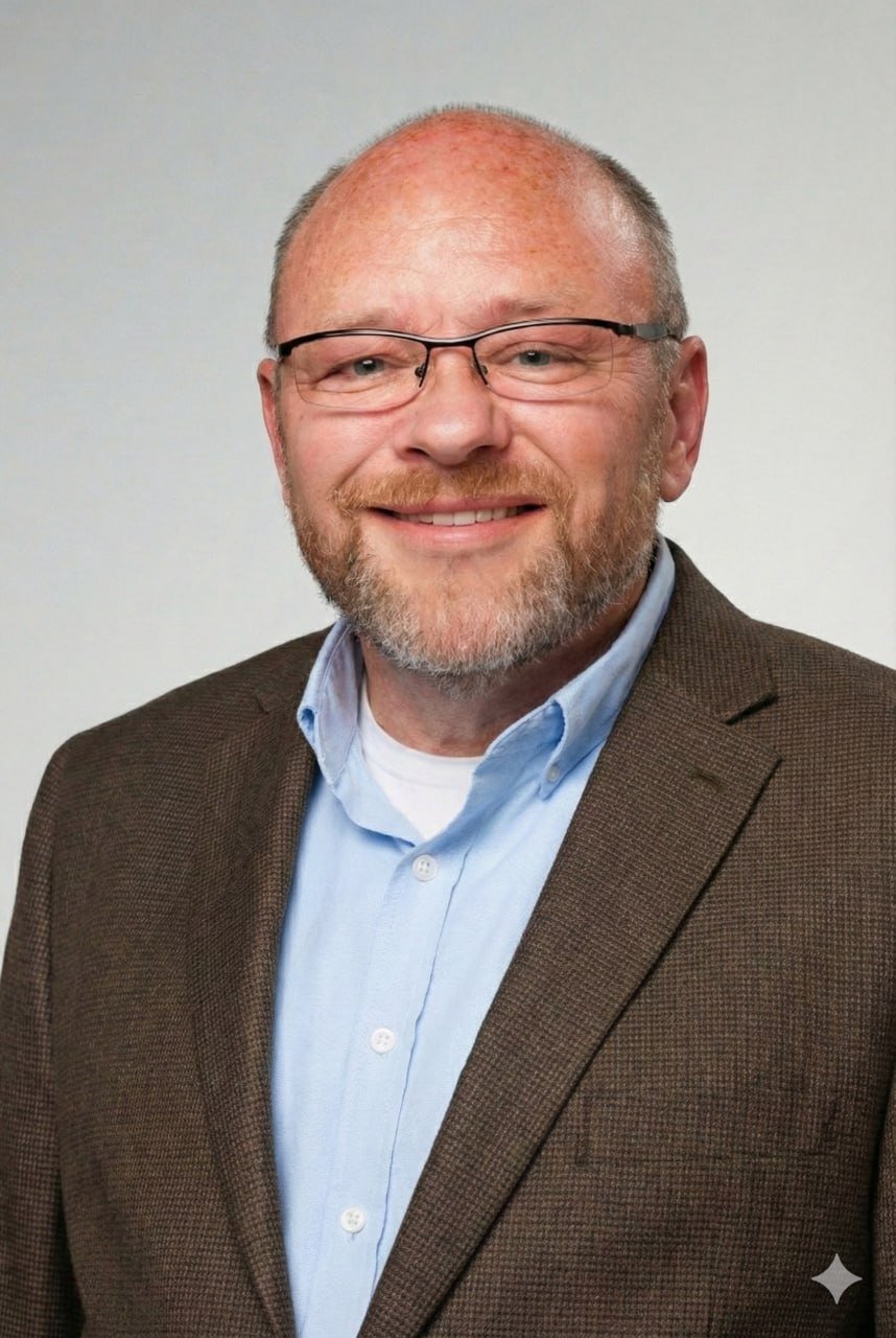 A smiling middle-aged man with glasses, a beard, and a bald head, wearing a brown blazer and light blue collared shirt, against a plain light background.