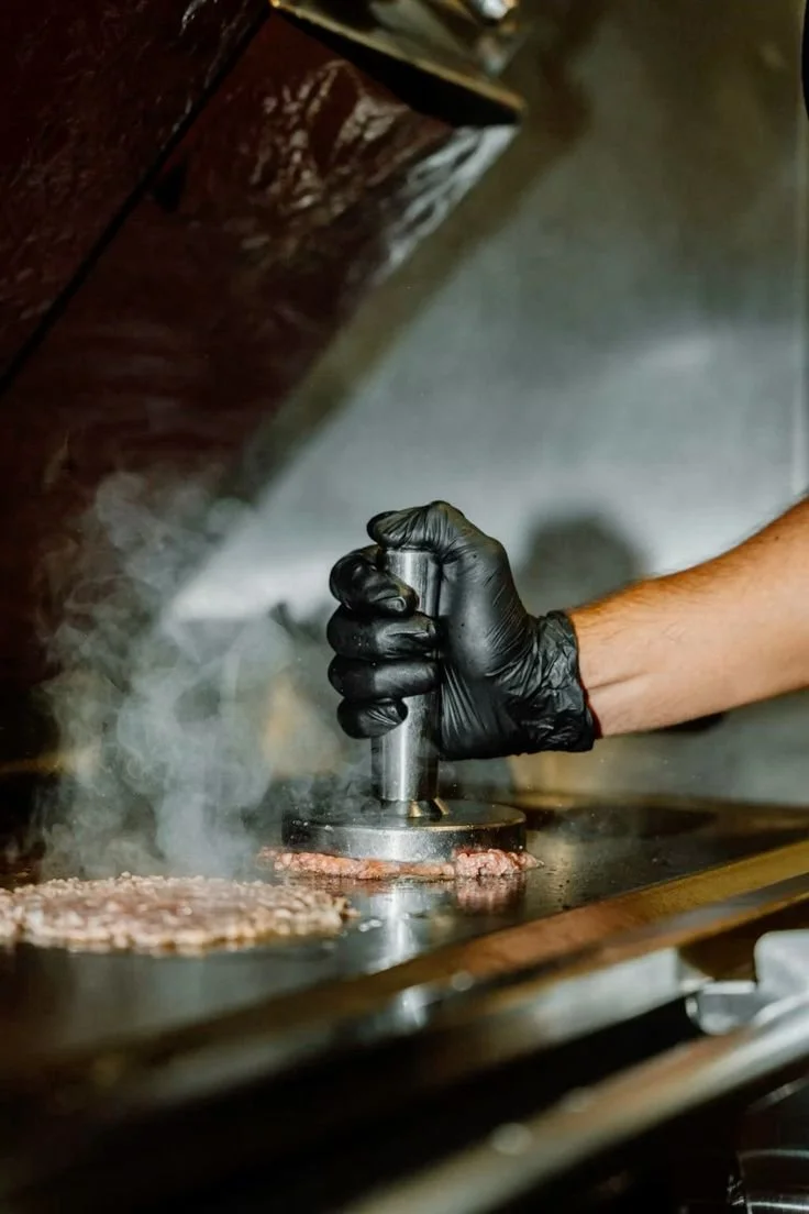 Person wearing a black glove pressing down on a burger patty on a flat top griddle, with steam rising.