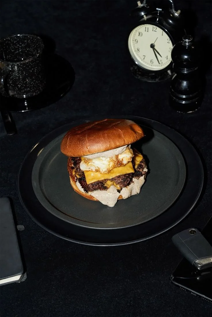 A cheeseburger with fried eggs in a brioche bun on a black plate, with a vintage clock and pepper grinder on a black table in the background.