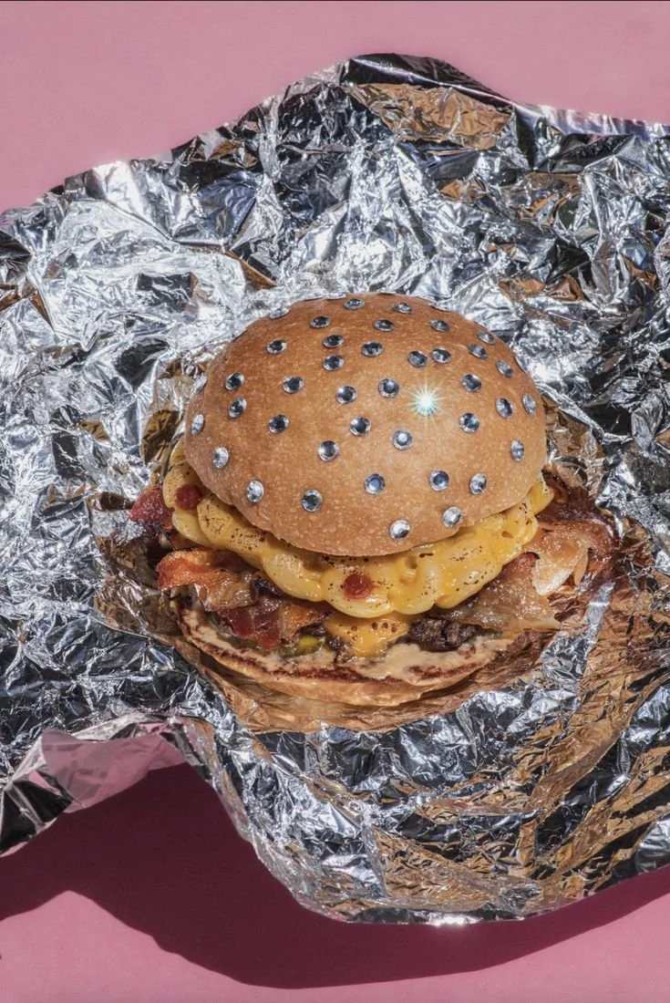 A cheeseburger with bacon and toppings in a sesame seed bun, wrapped in crinkled aluminum foil on a pink background.