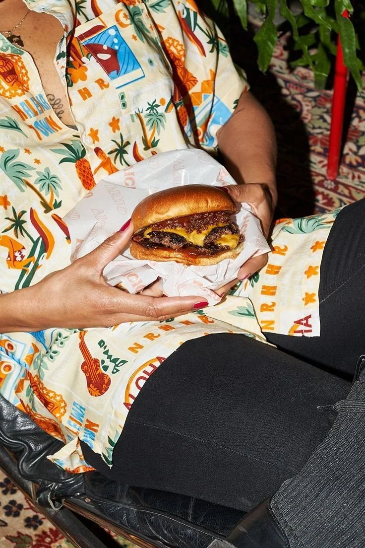 Person holding a cheeseburger with beef patty and melted cheese, seated with colorful tropical-themed clothing, on a patterned rug.