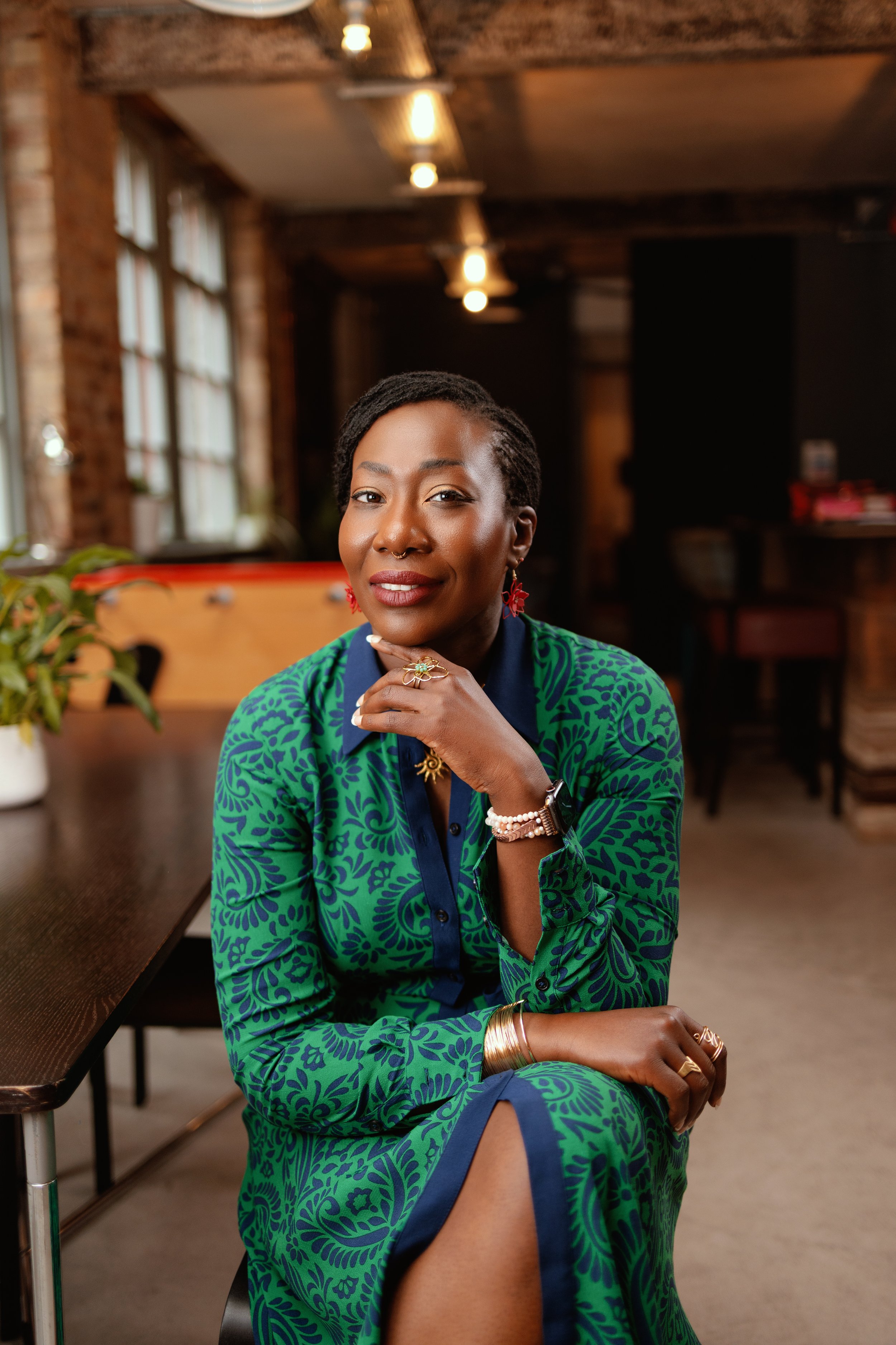 A woman with short black hair, wearing a green and blue patterned dress, sitting indoors with a confident smile, surrounded by exposed brick walls and large windows.