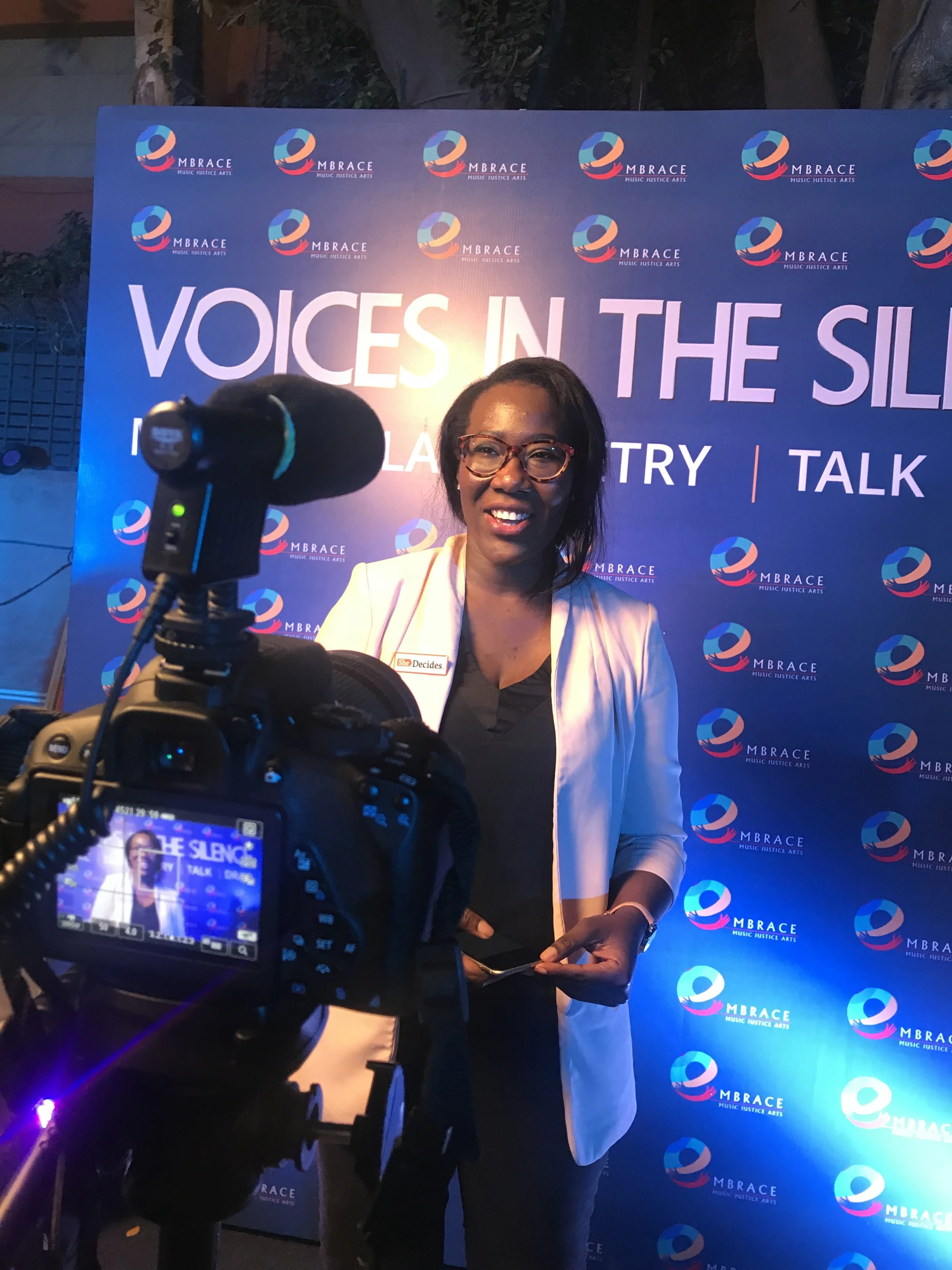 Woman with glasses and a white blazer speaking in front of a blue backdrop with event branding, being filmed or photographed.