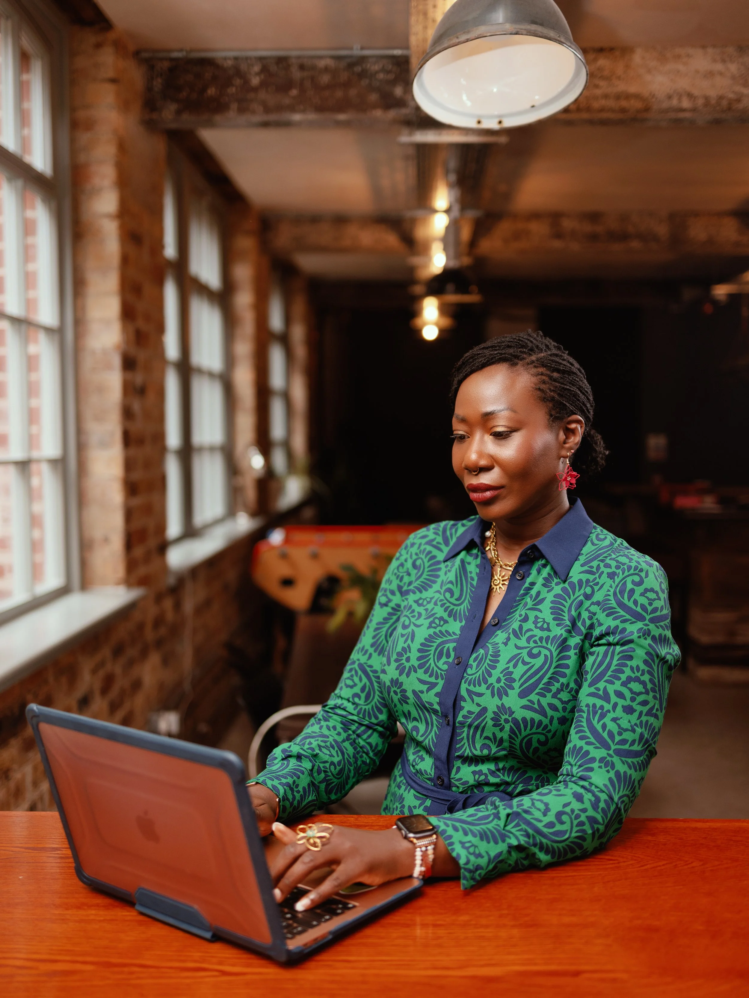 A woman with short dreadlocks using a laptop in a cafe with exposed brick walls and large windows.