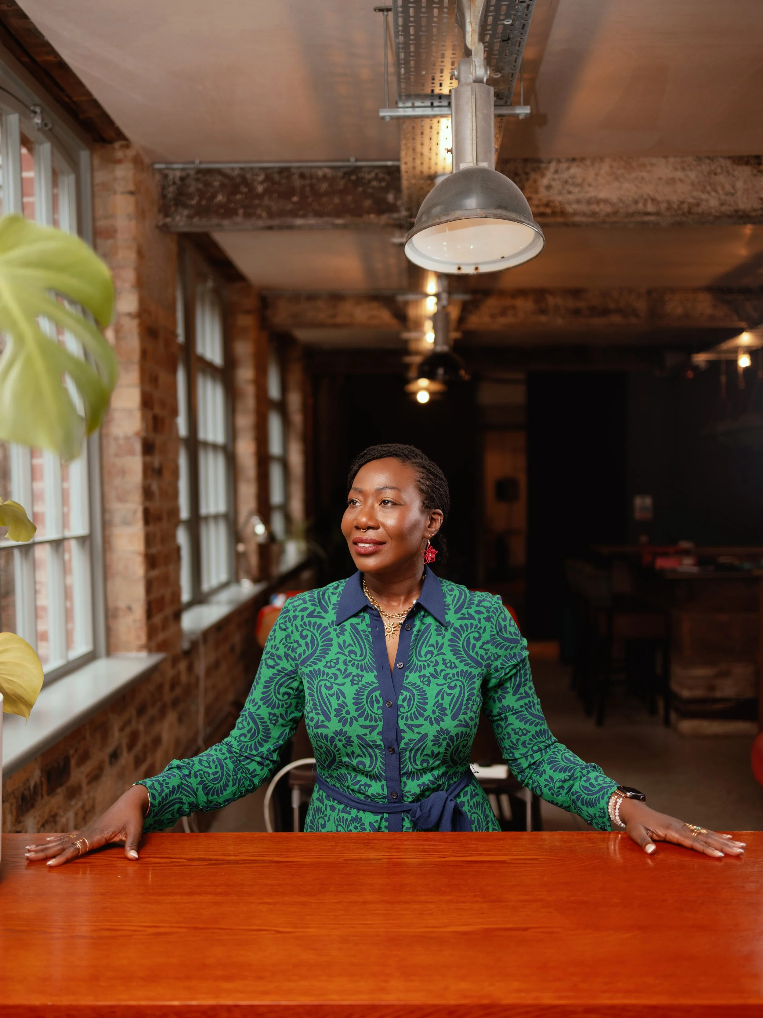 Dr Kizanne James Woman in green and blue patterned dress standing behind a wooden table in a rustic, industrial-style restaurant or cafe.