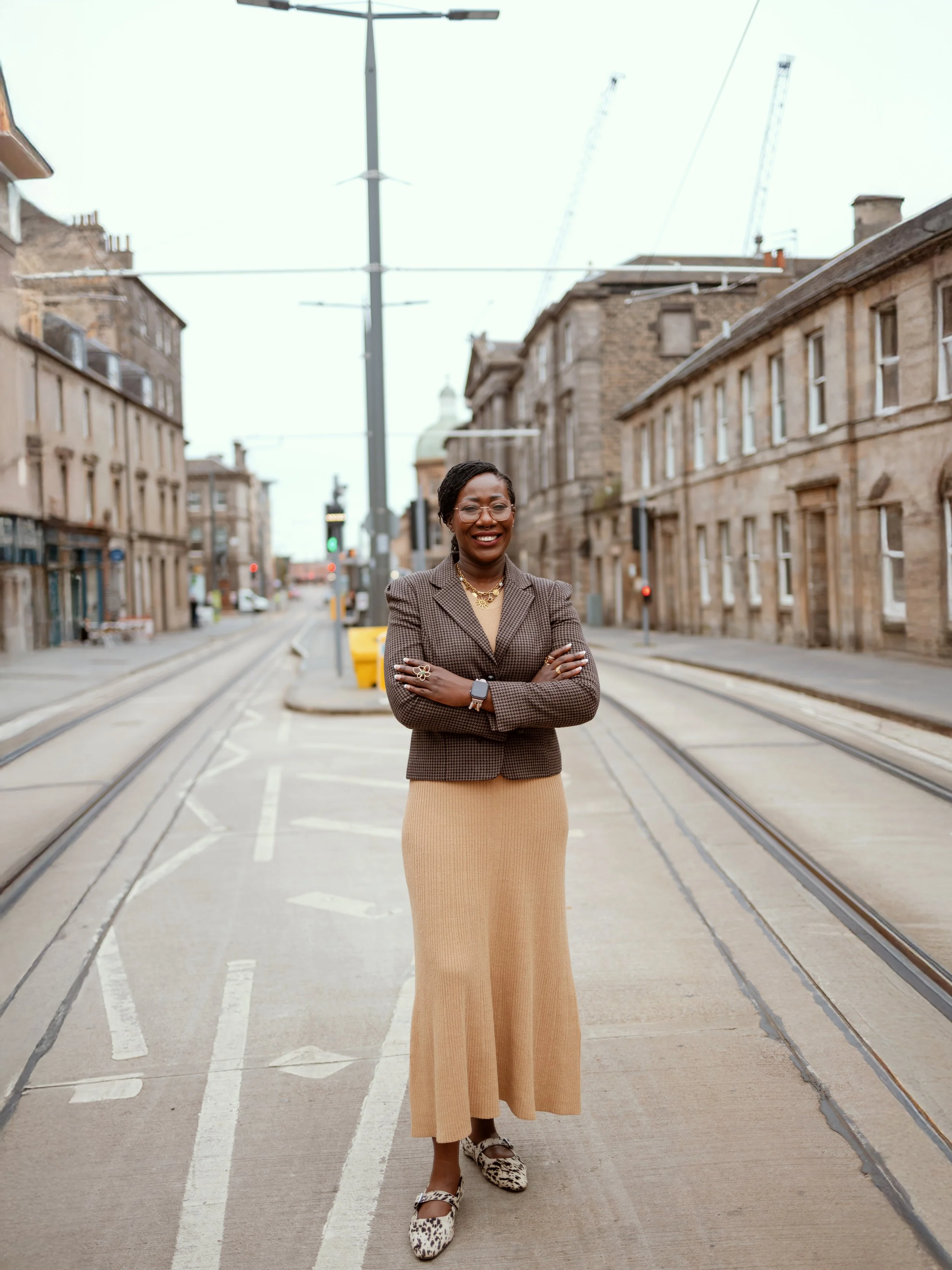 Dr Kizanne James A woman in business attire standing confidently on a city street with tram tracks, old buildings, and crane construction in the background.