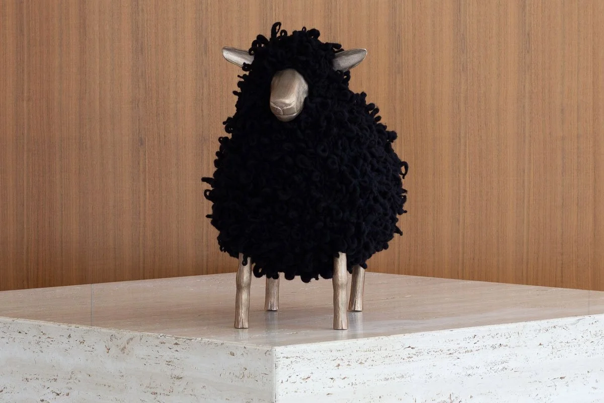 A black knitted sheep footstool with bronze legs, a bronze face, and bronze ears stands on a marble plinth in front of a dark wooden wall.