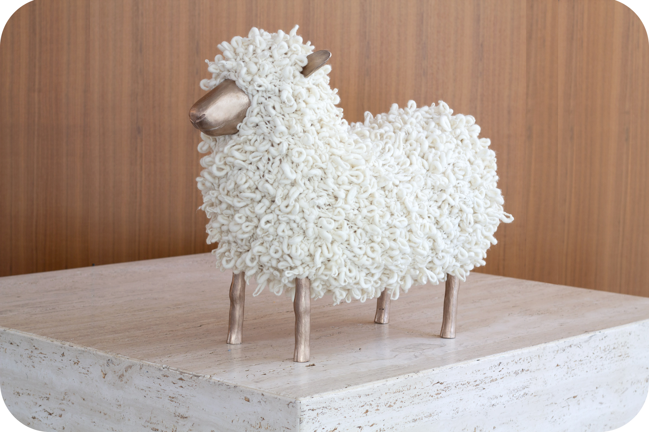 A white knitted sheep footstool with bronze legs, a bronze face, and bronze ears stands on a marble plinth in front of a dark wooden wall.