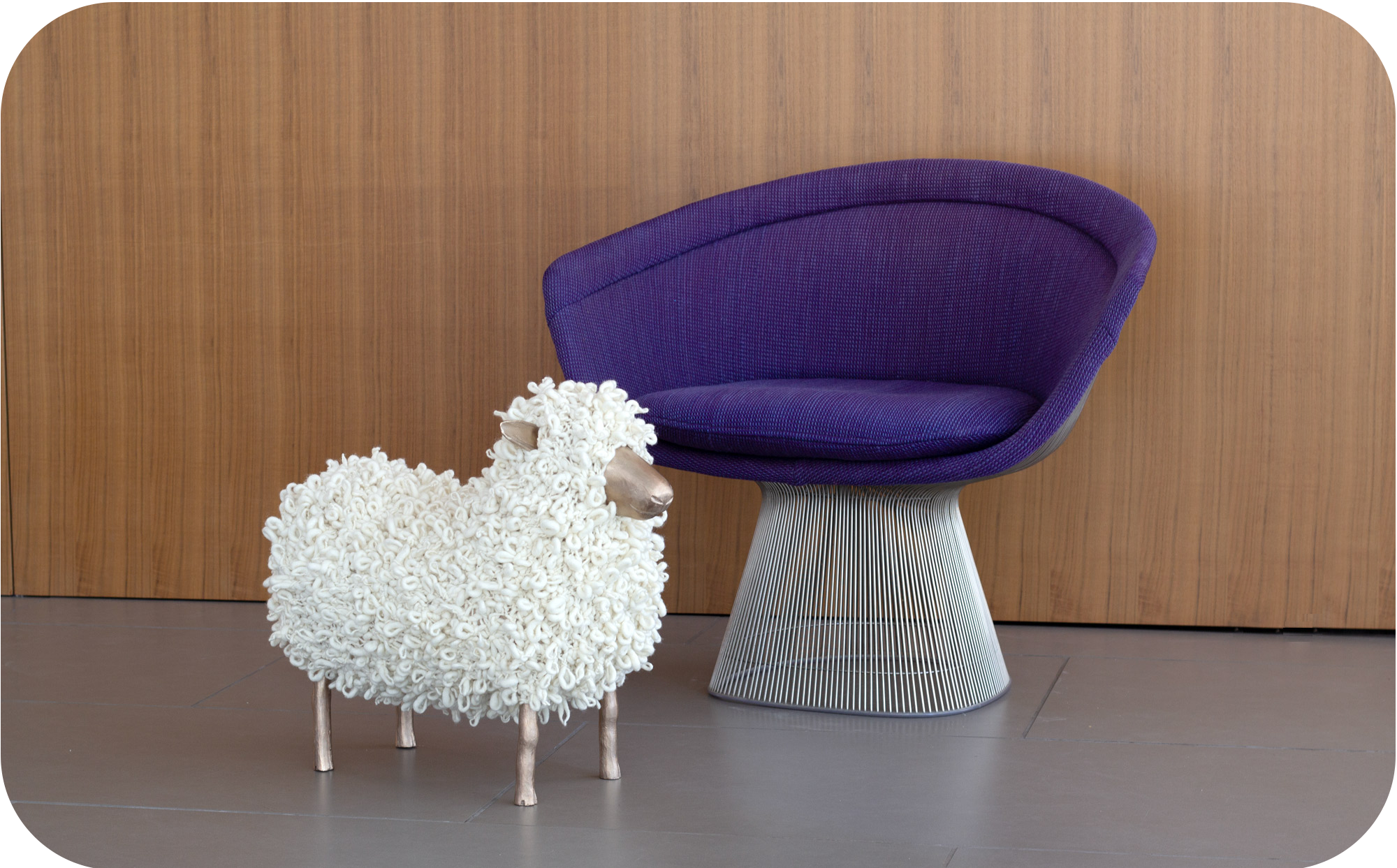 A white knitted sheep with bronze legs, face, and ears stands in front of a purple 1960s chair and a wooden wall