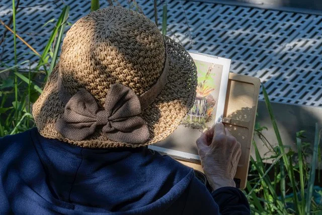 A person wearing a wide-brimmed straw hat with a bow, sitting outdoors and using a tablet device surrounded by tall green grass.