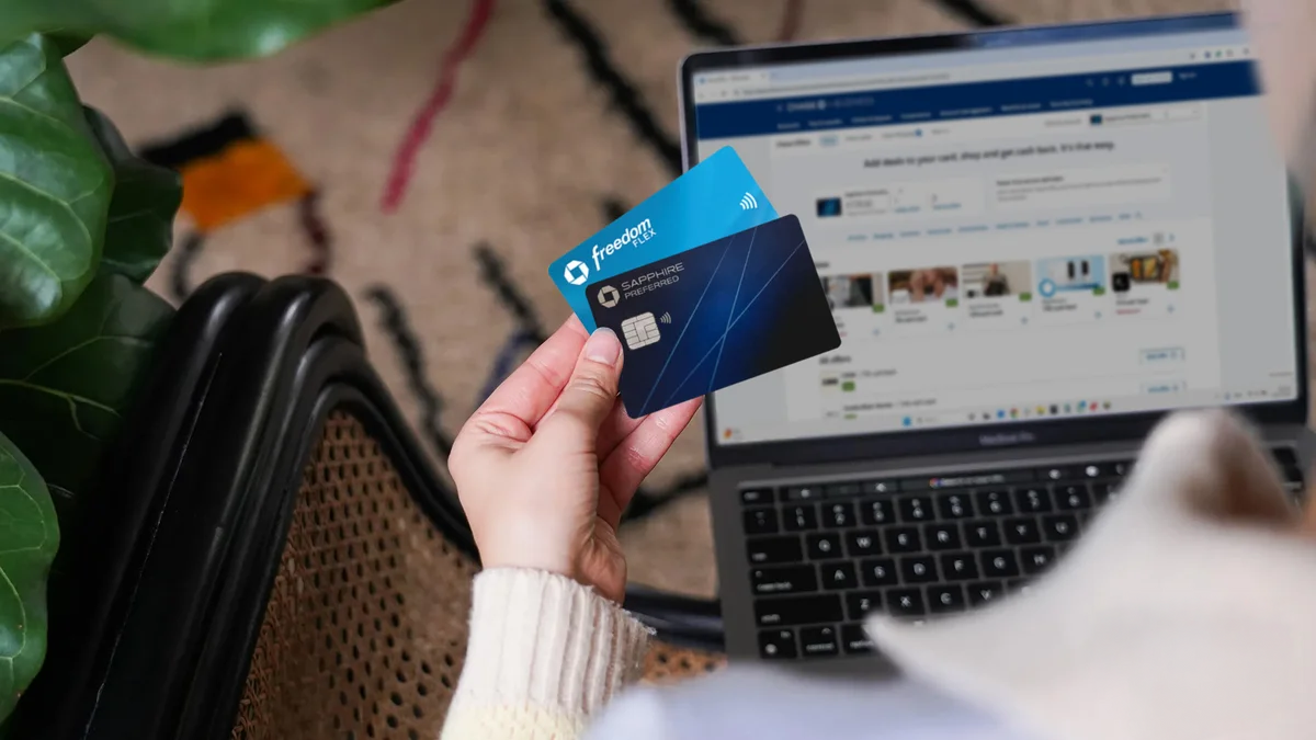 Person holding two credit cards with a laptop open to a social media page in the background.