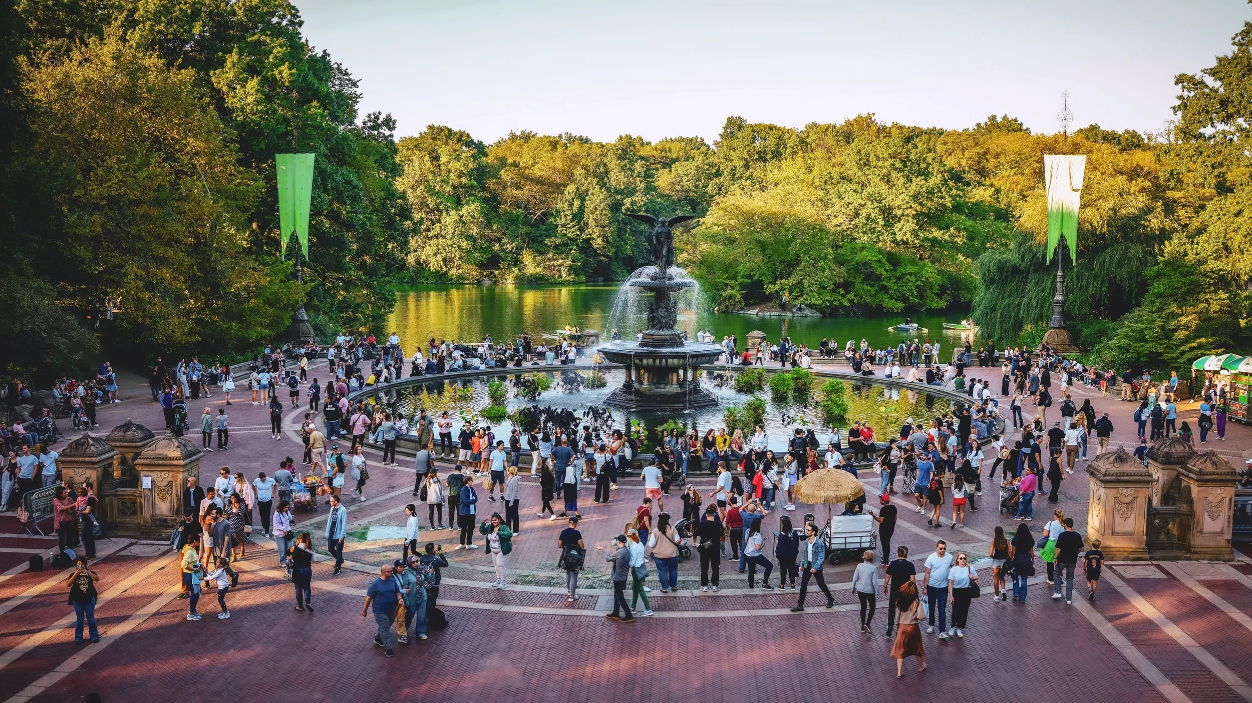 Crowd of people gathered around a large fountain in a park with trees and a lake in the background.