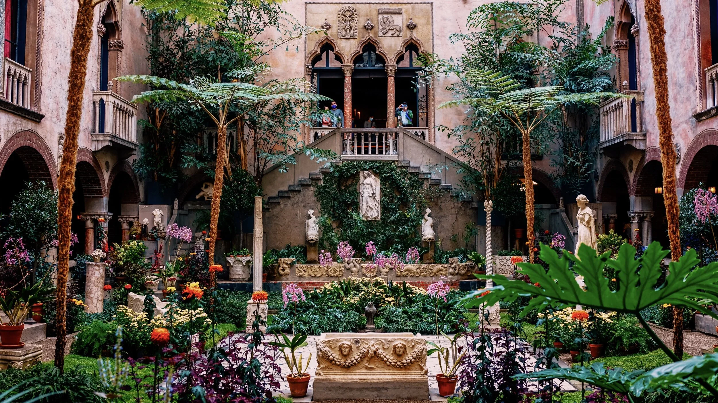 Indoor courtyard filled with lush green plants, colorful flowers, and classical sculptures, with stairs towards an open balcony at the top center. The courtyard is framed by pink walls with arches and decorative carvings.