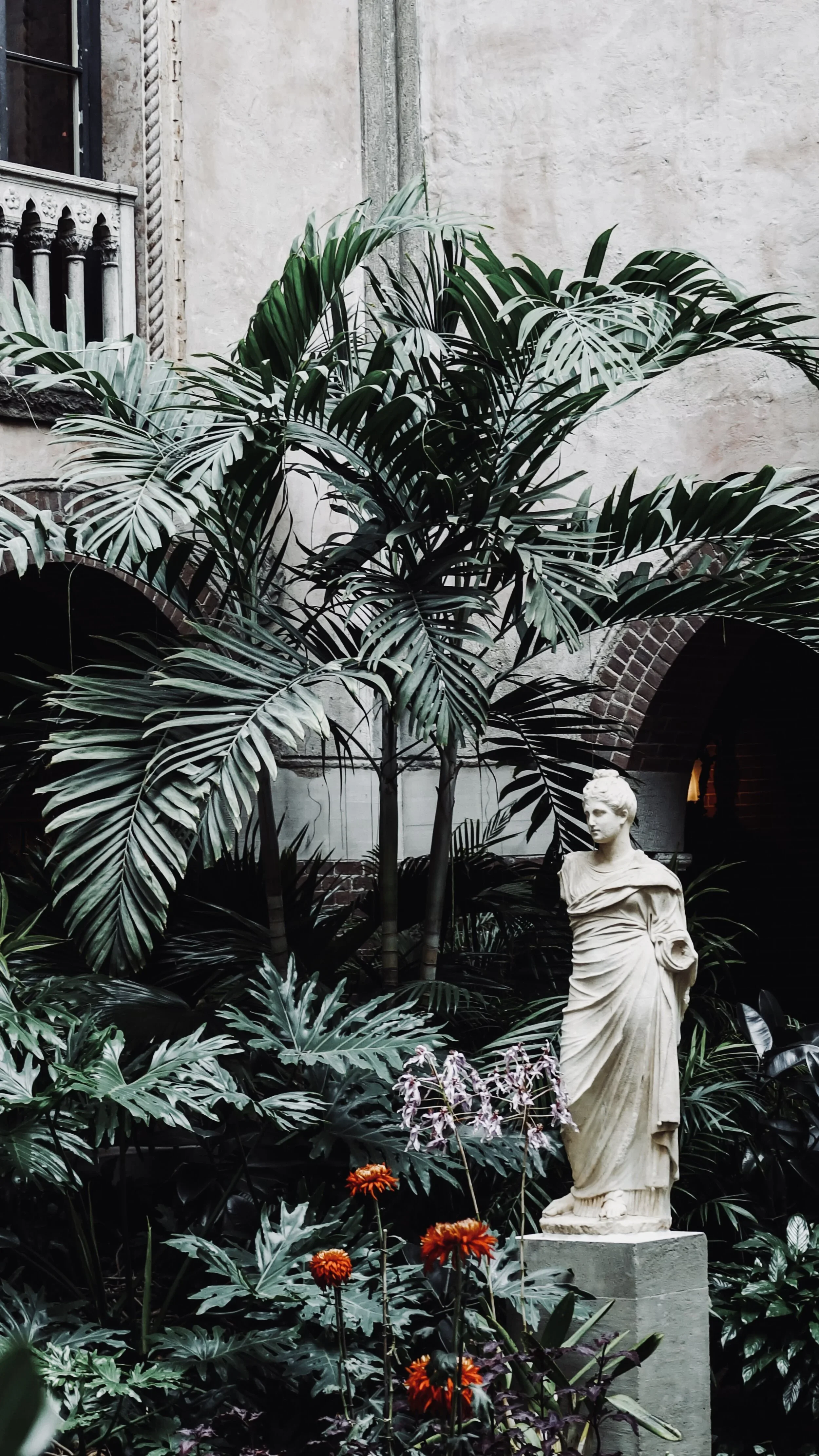 A classical statue of a robed woman in a lush indoor garden with large green tropical plants and bright orange flowers.