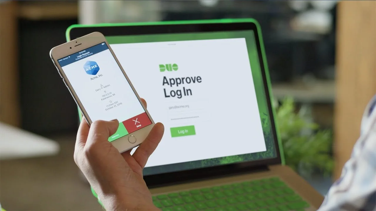 Person holding a smartphone with a registration or login screen for Acme Inc., featuring approval and denial buttons, in front of a laptop displaying an approval login page for a different service.