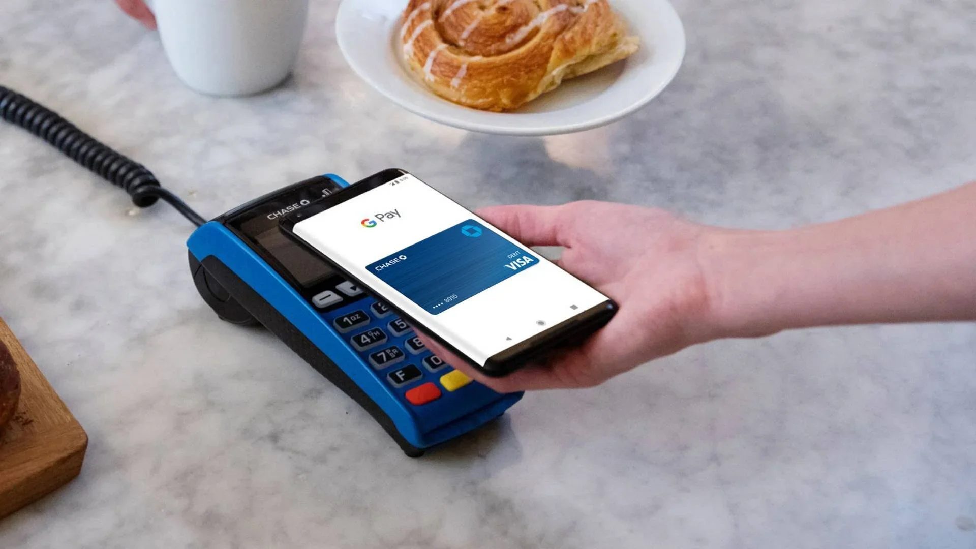 A person using a smartphone to make a contactless payment with Google Pay on a point-of-sale terminal, with a plate of a croissant and a cup in the background.