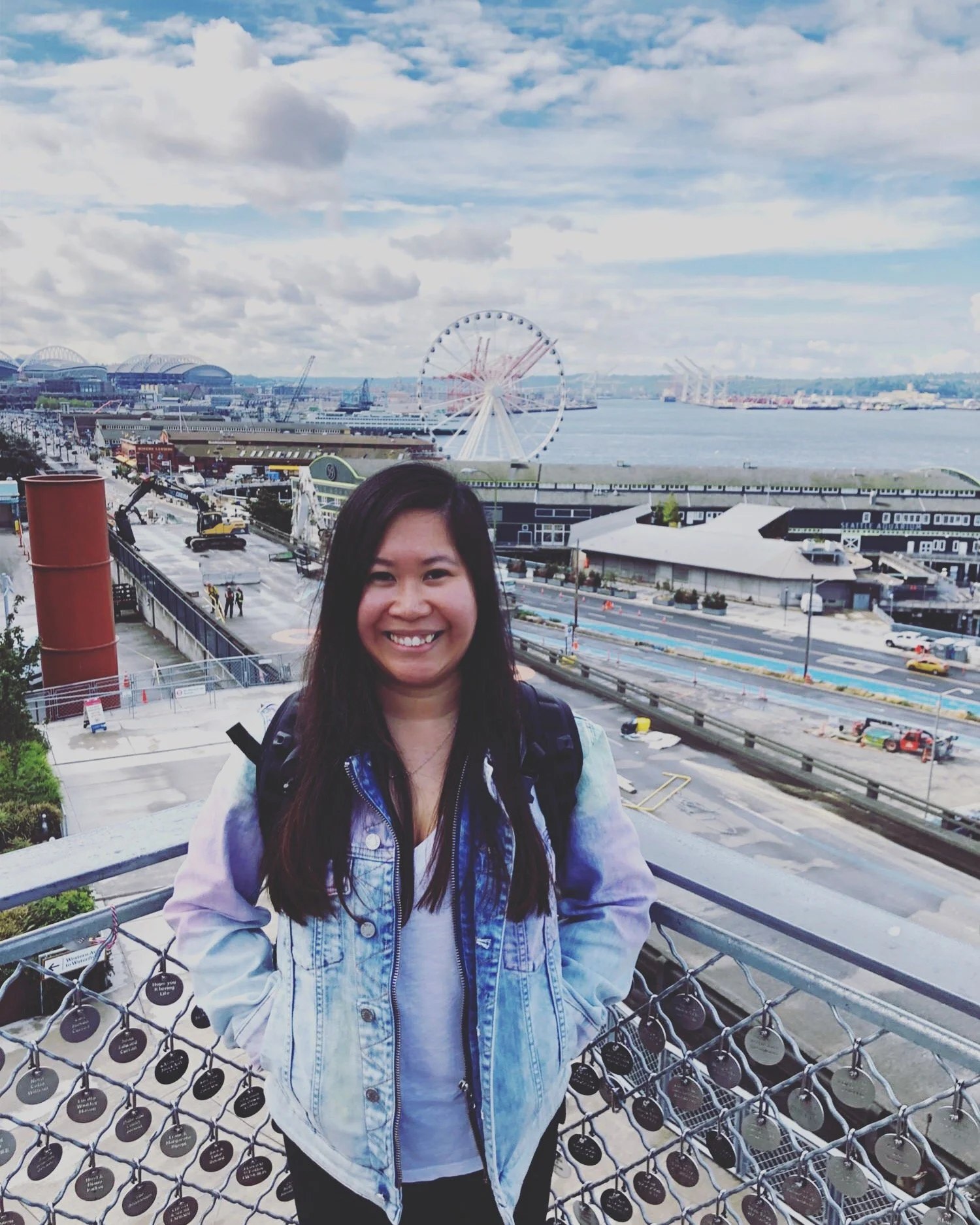 A woman smiling while standing outdoors near a waterfront with a Ferris wheel, water, and cityscape in the background.