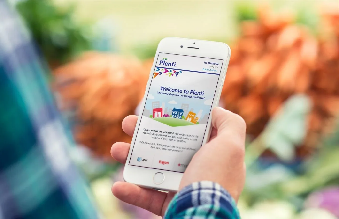 Person holding a smartphone displaying the Plenti rewards app, with a blurred indoor or outdoor background.