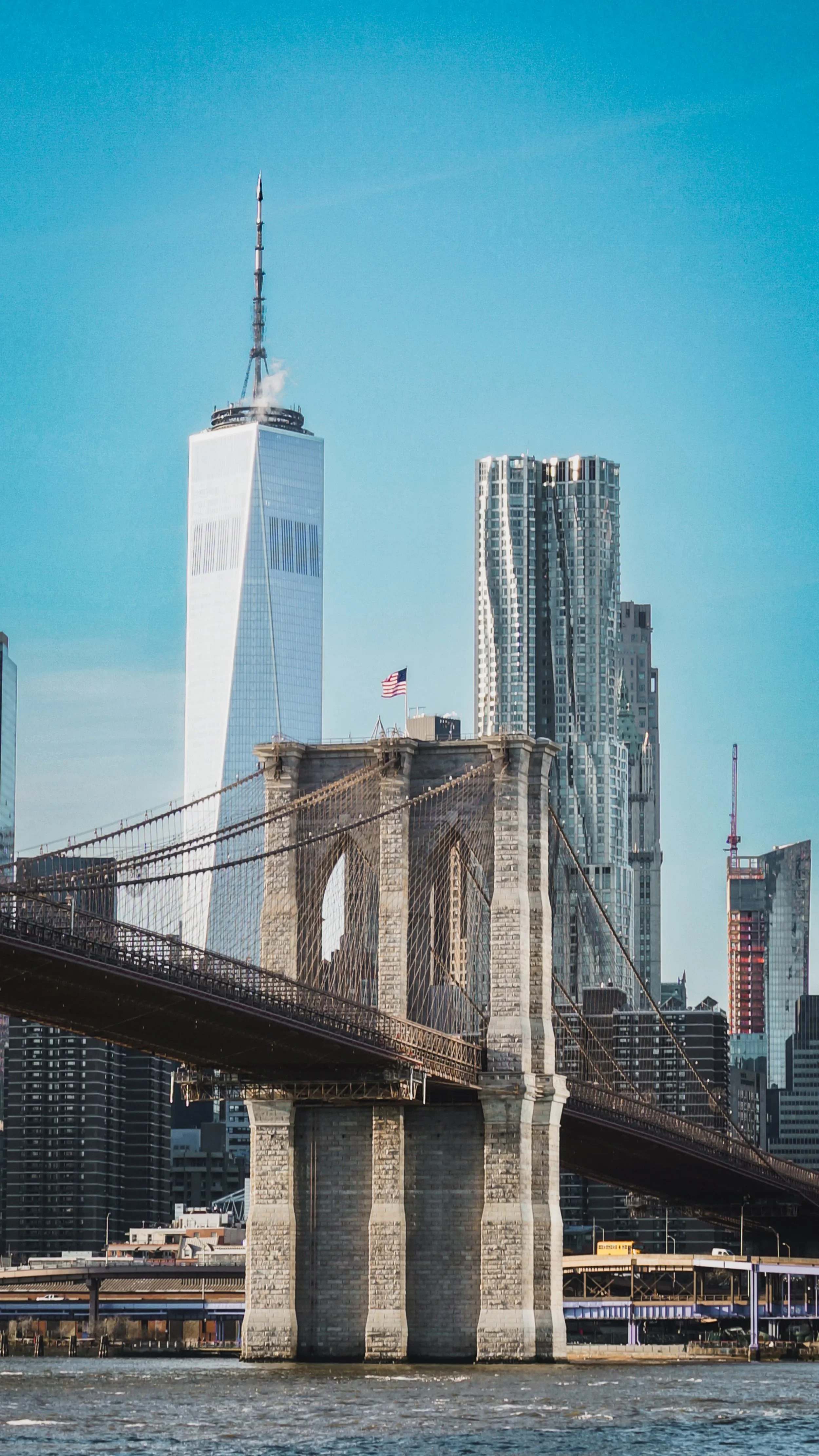 View of the Brooklyn Bridge with the Manhattan skyline in the background, including the One World Trade Center and various skyscrapers, under a clear blue sky.