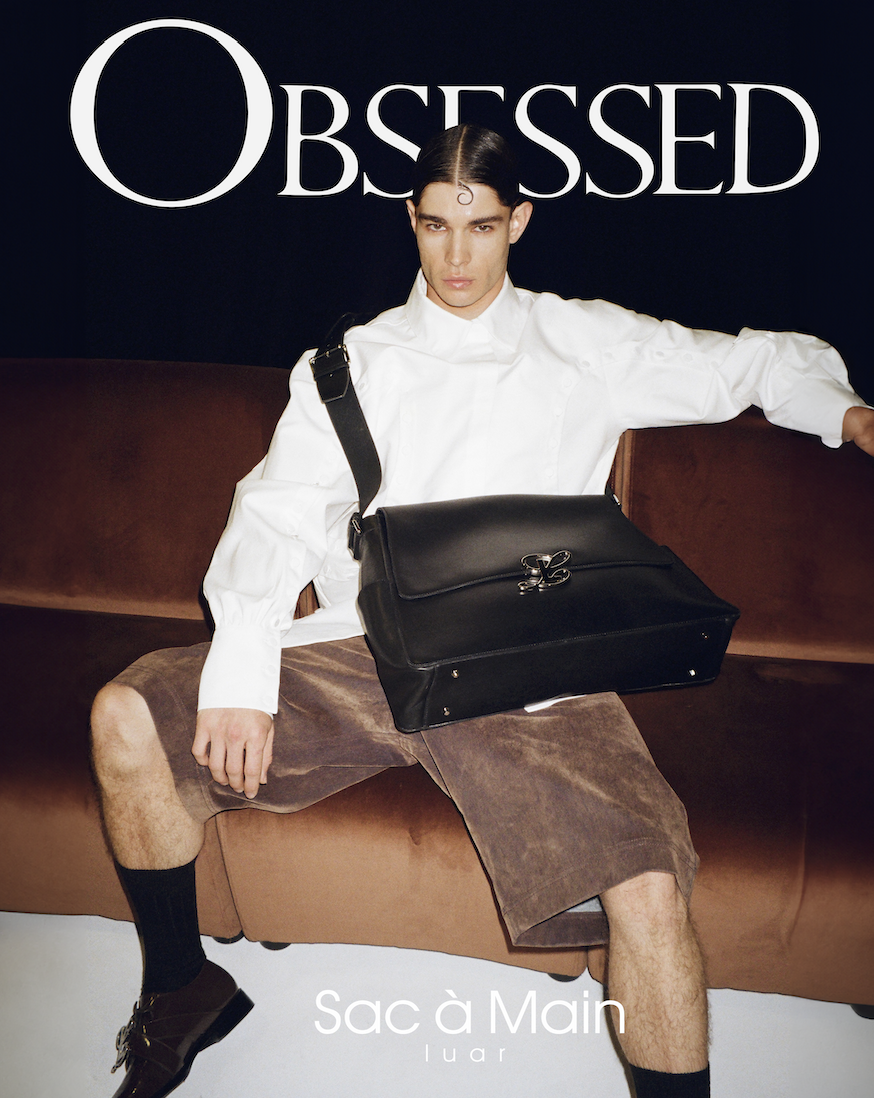 A young man with dark, slicked-back hair sitting on a brown couch, wearing a white shirt and brown shorts, holding a large black leather briefcase, with the word 'OBSESSED' in large white letters above him and 'Sac à Main' in smaller white text at the bottom.