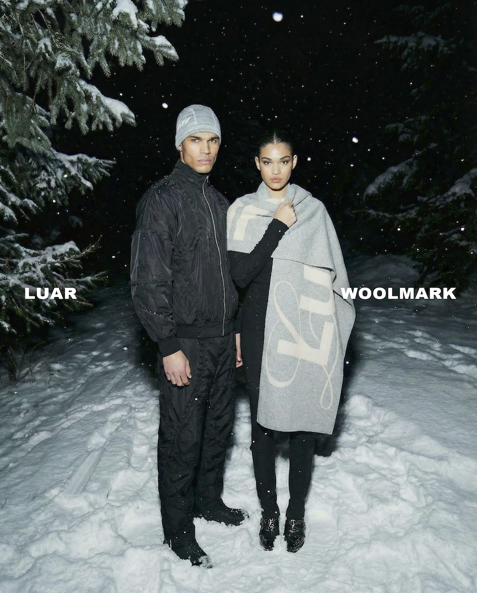 Two models standing outdoors in the snow at night. The male model is dressed in a black outfit with a gray beanie, and the female model is wrapped in a large gray scarf with a design on it. Tall pine trees with snow on their branches are in the background. The words 'LUAR' and 'WOOLMARK' are written on the image.