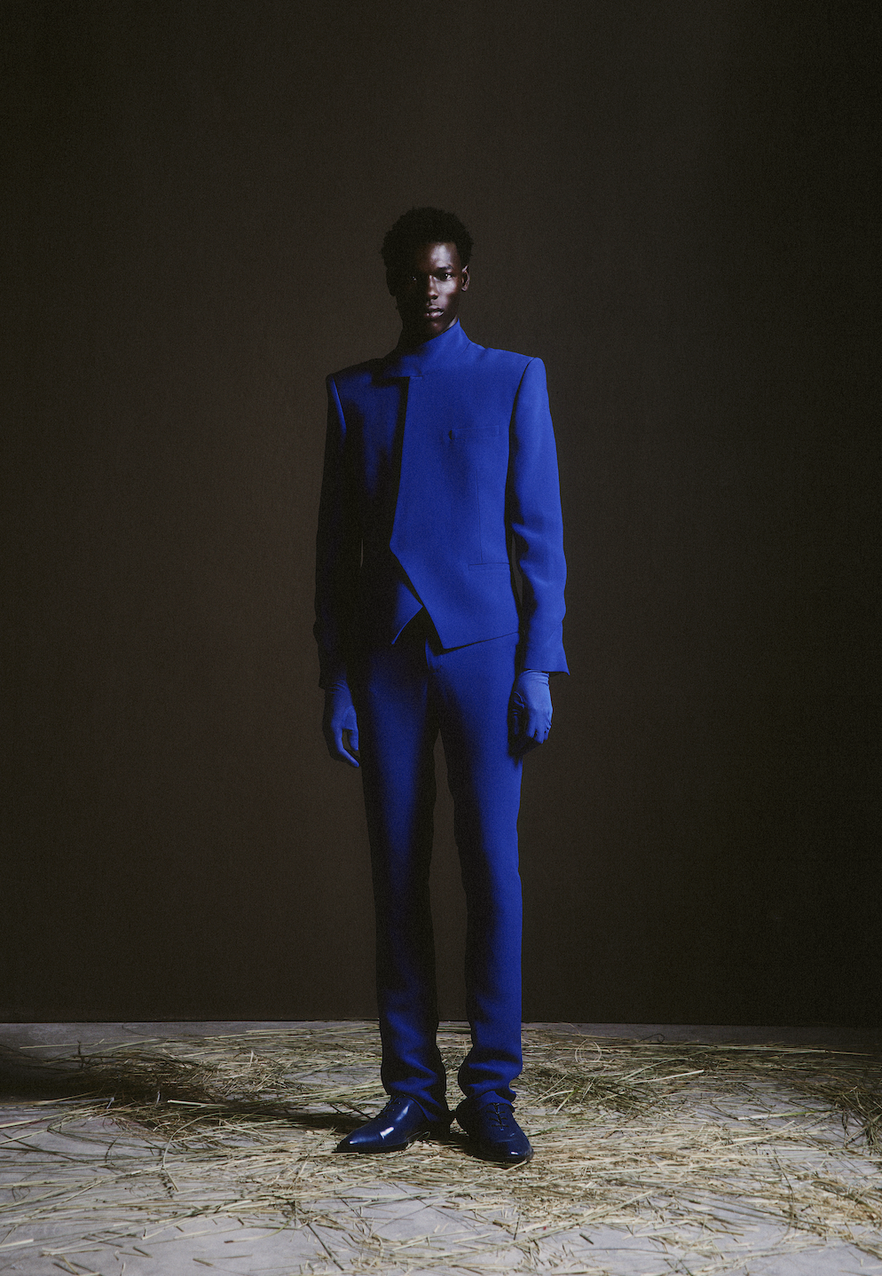 A person with dark skin dressed in a royal blue suit and gloves, standing on a floor covered with straw against a dark background.