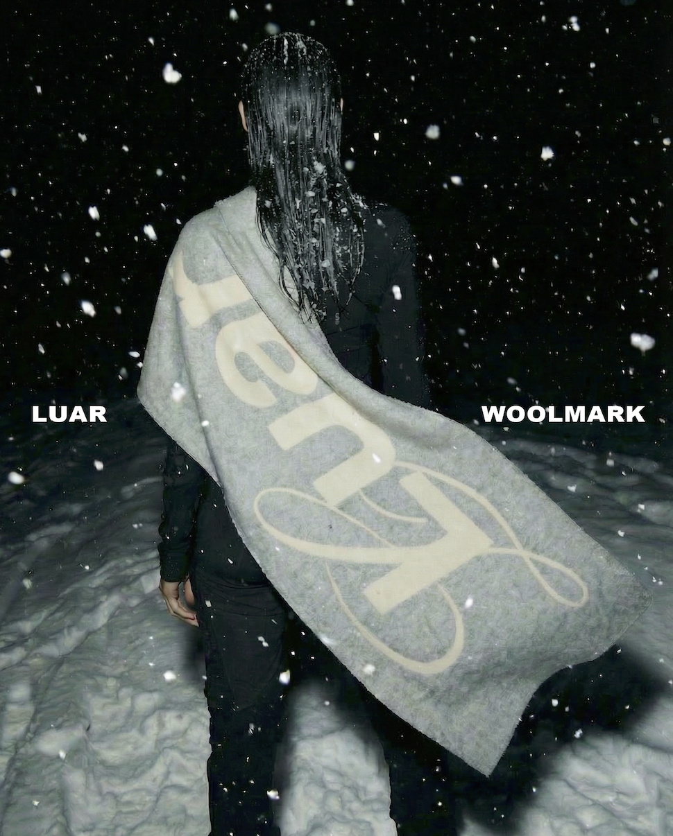 Person standing outdoors at night in falling snow, wearing black clothing, with long wet hair covering their face, holding a large gray Woolmark blanket or flag.