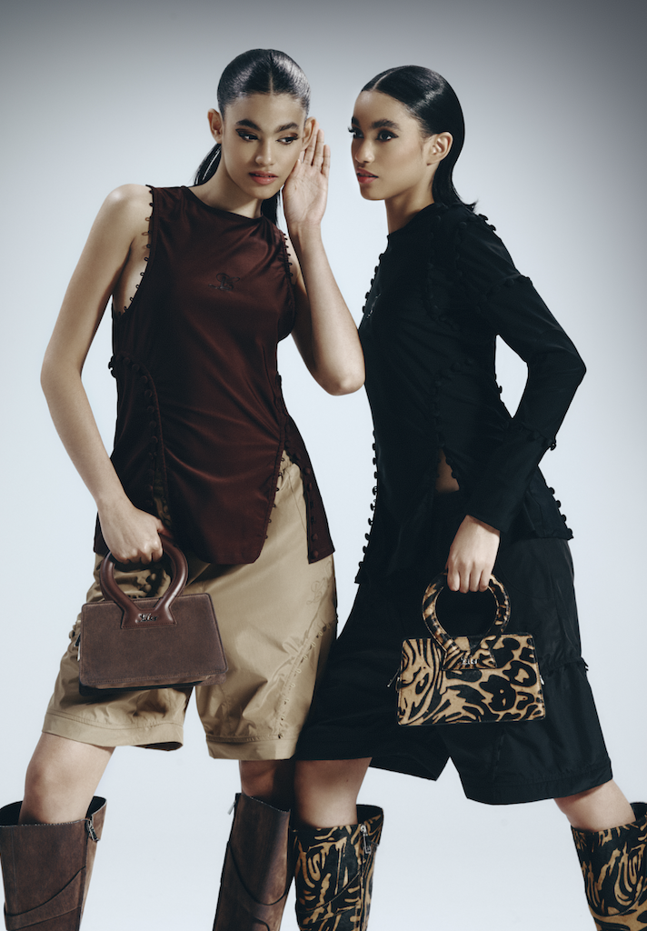 Two women with long dark hair and sleek hairstyles posing in fashion-forward outfits, holding stylish handbags and wearing tall boots, against a plain gray backdrop.