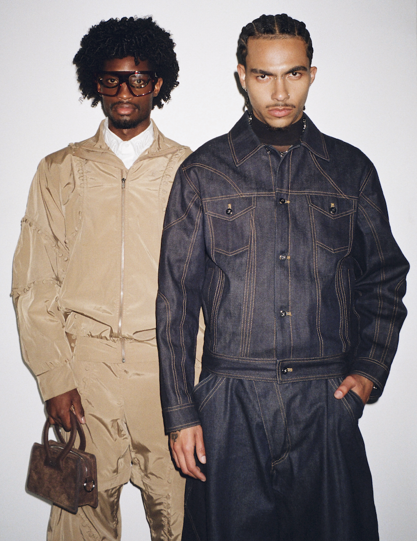 Two young men posing against a plain white background, one wearing a beige tracksuit with glasses and holding a brown purse, the other wearing a dark denim jacket and matching pants.