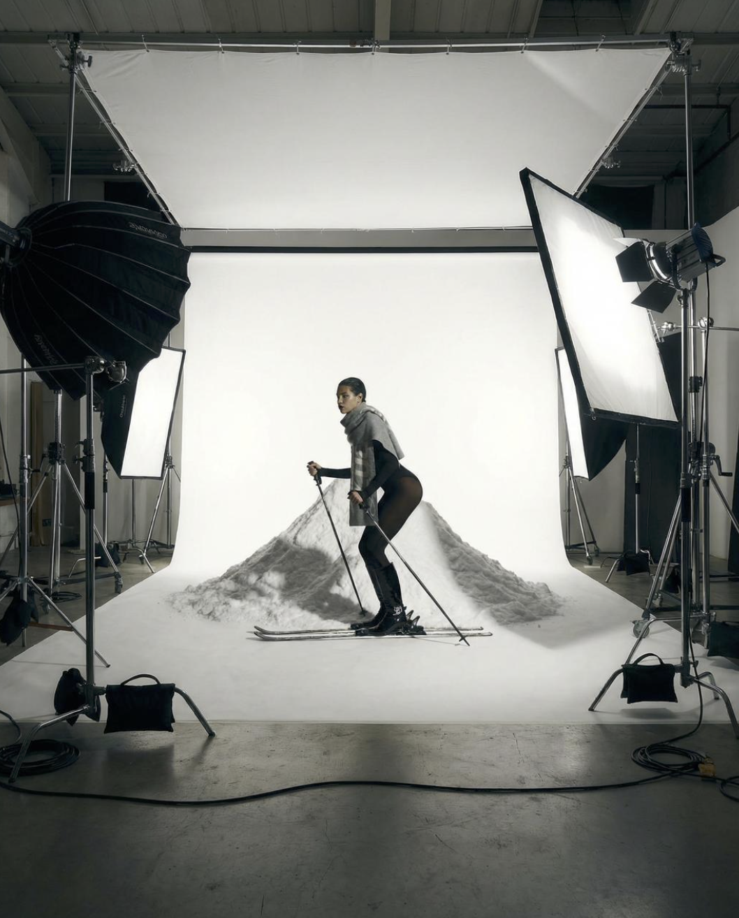 A woman in winter clothing and skis standing on snow in a photography studio with professional lights and a white backdrop.