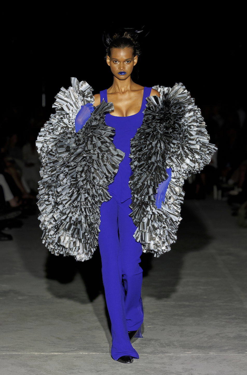 Model walking down runway in cobalt blue jumpsuit with large silver ruffle coat, purple lipstick, and matching purple glove on right hand.
