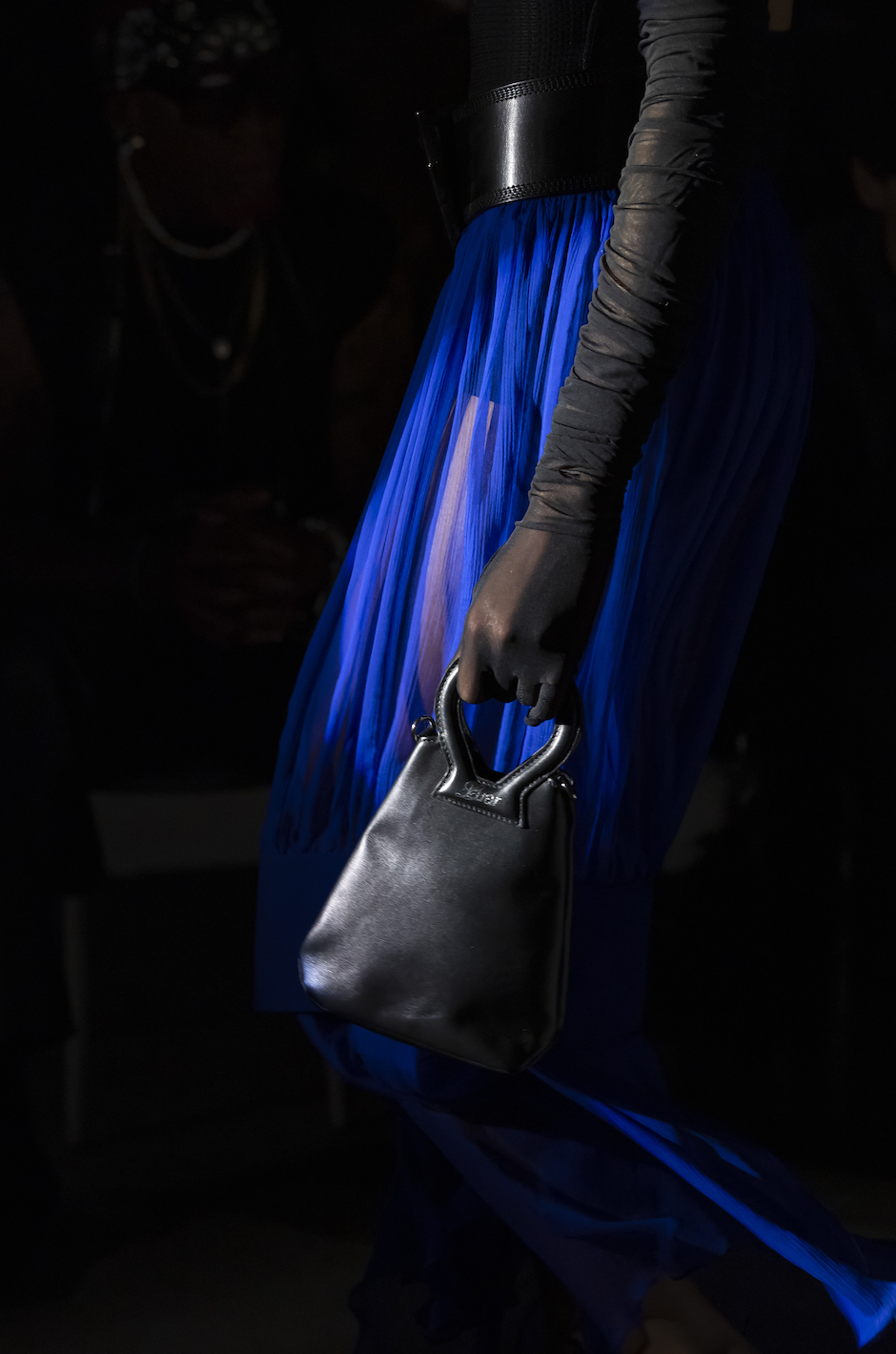 Close-up of a person wearing a black top with long sleeve gloves, holding a small black handbag, and wearing a blue pleated skirt in a dark setting.