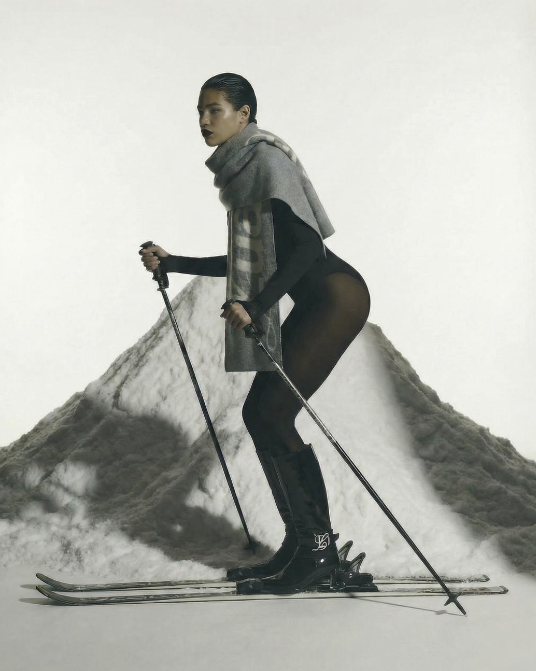 Fashion model wearing a black bodysuit, black boots, and a gray scarf, standing on skis with ski poles, in front of a mountain of snow.